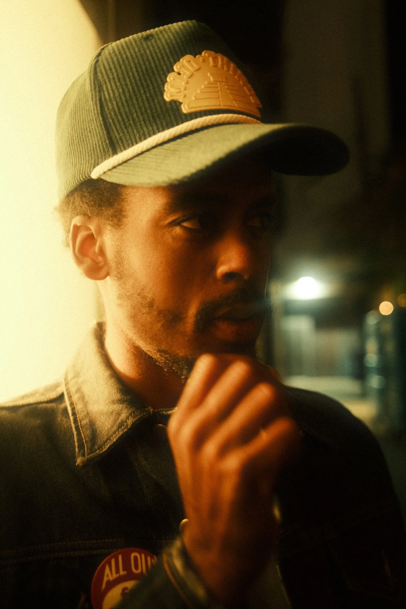A young man wearing a green corduroy baseball cap with a patch and a dark jacket is looking toward the camera, with his hand near his face. The background is blurry with some lights.