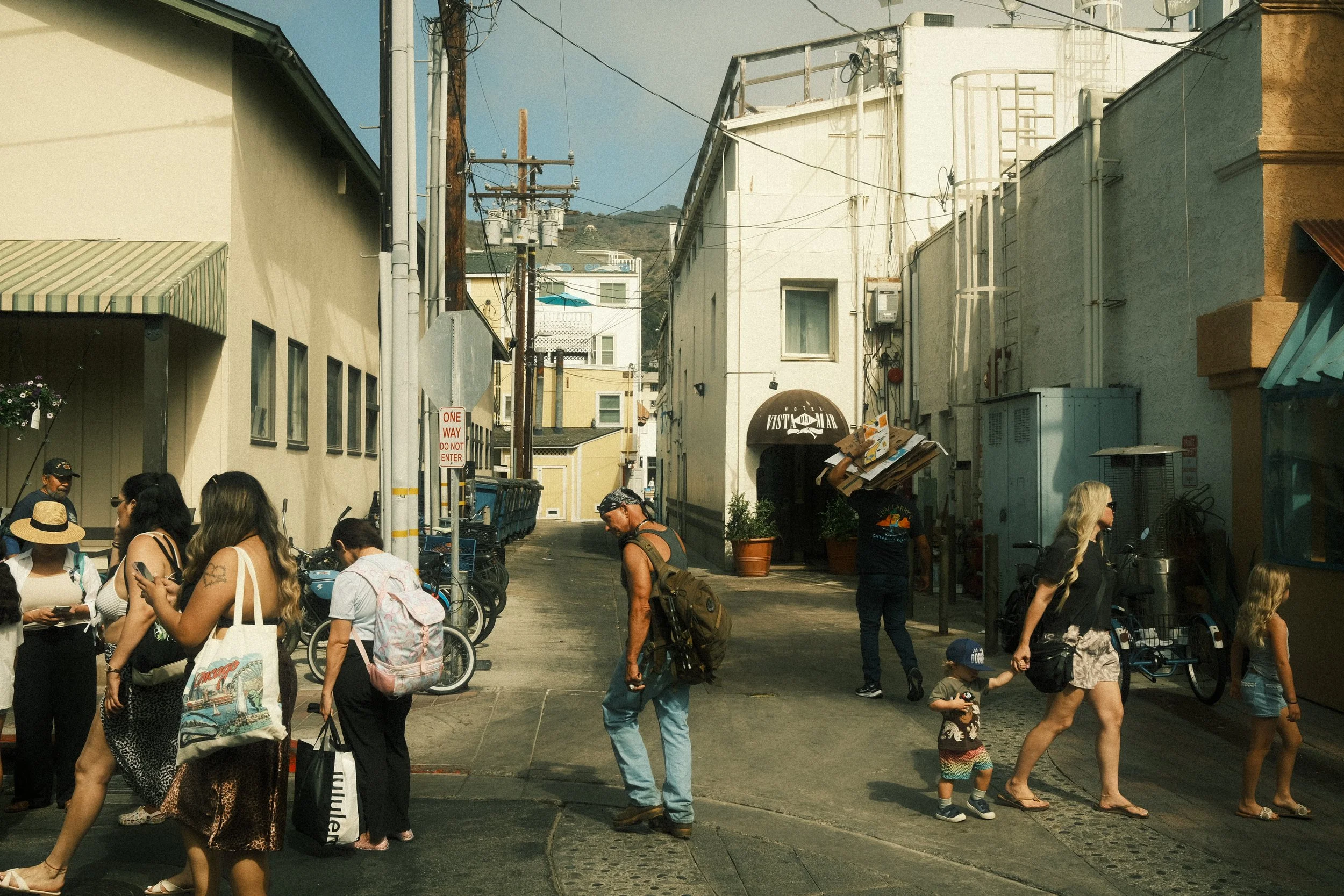 A busy alleyway with people walking and standing, some using phones. Buildings line each side of the alley, with utility poles and wires overhead. There is a sign that reads 'One Way Do Not Enter' and a storefront with a sign that says 'VISTA MAR.' T