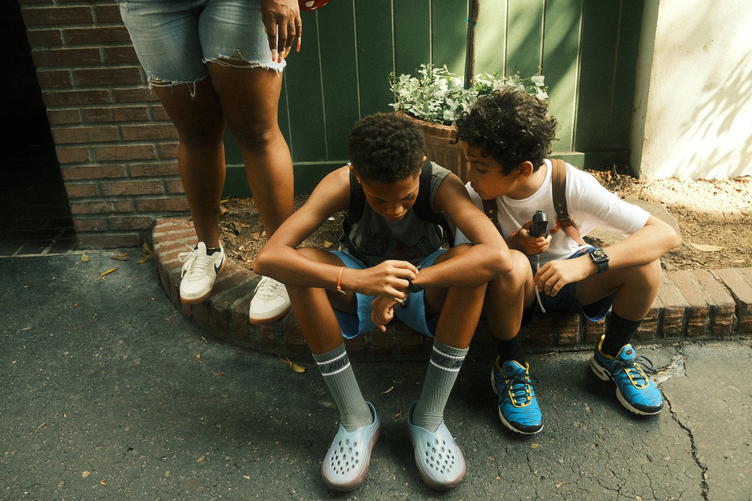 Two boys sitting on a low brick wall, one holding a portable fan, while a woman stands nearby wearing ripped shorts and sneakers. There are plants and a green fence in the background.