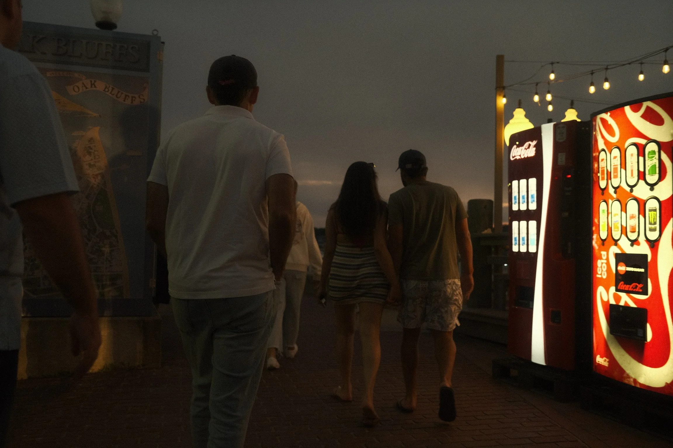 People standing in line at night near illuminated vending machines with string lights overhead.