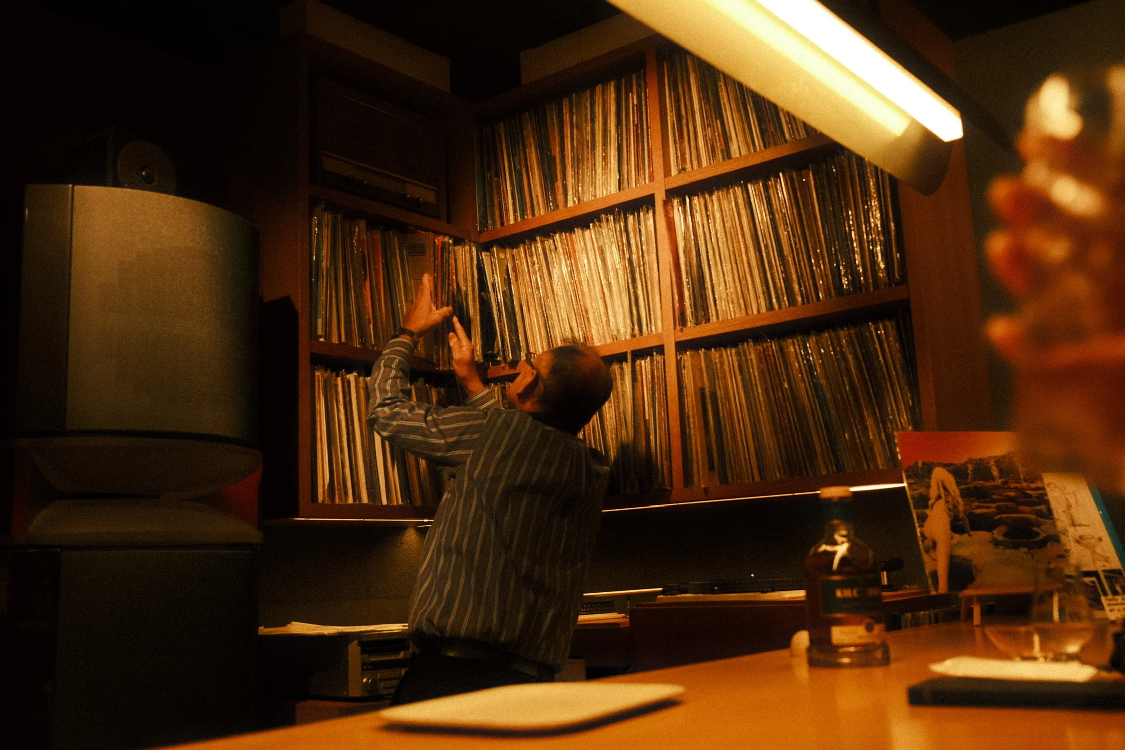 A man in a striped shirt reaching up towards a large bookshelf filled with vinyl records in a dimly lit room.