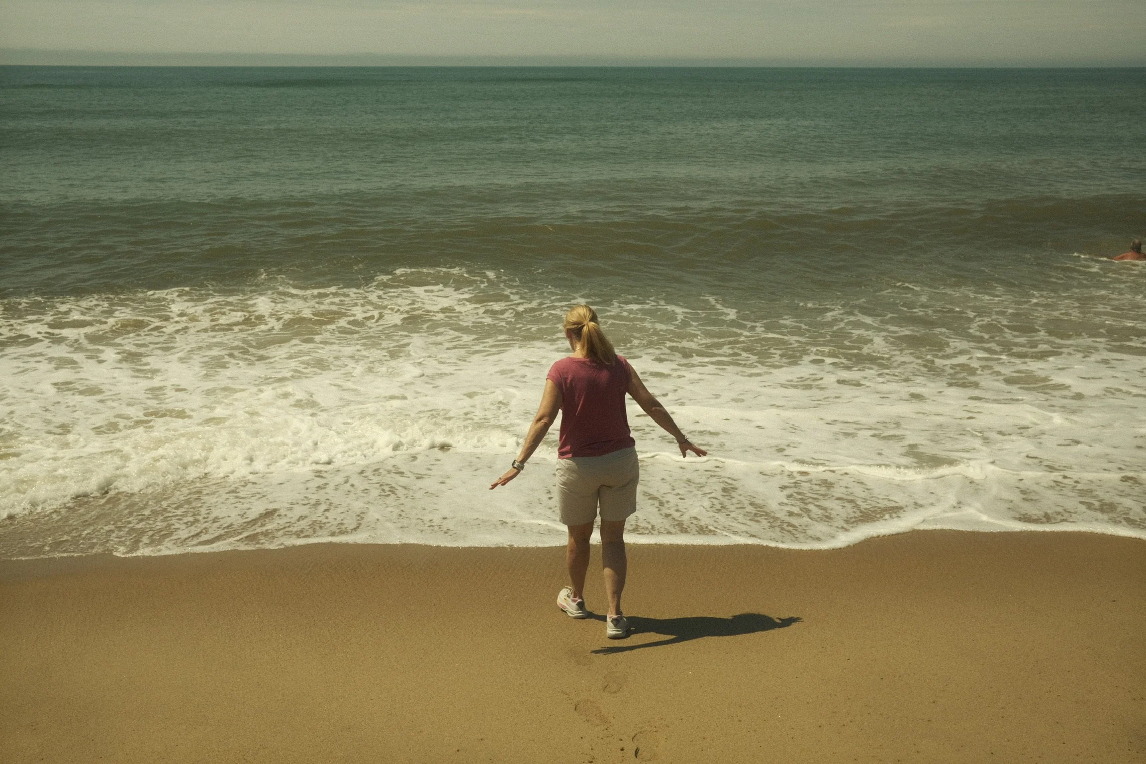 A woman with blonde hair tied back, wearing a pink shirt, beige shorts, and sneakers, is walking on the beach near the shoreline with her arms outstretched. The ocean waves are crashing onto the sand, and there is a man swimming in the water to the r