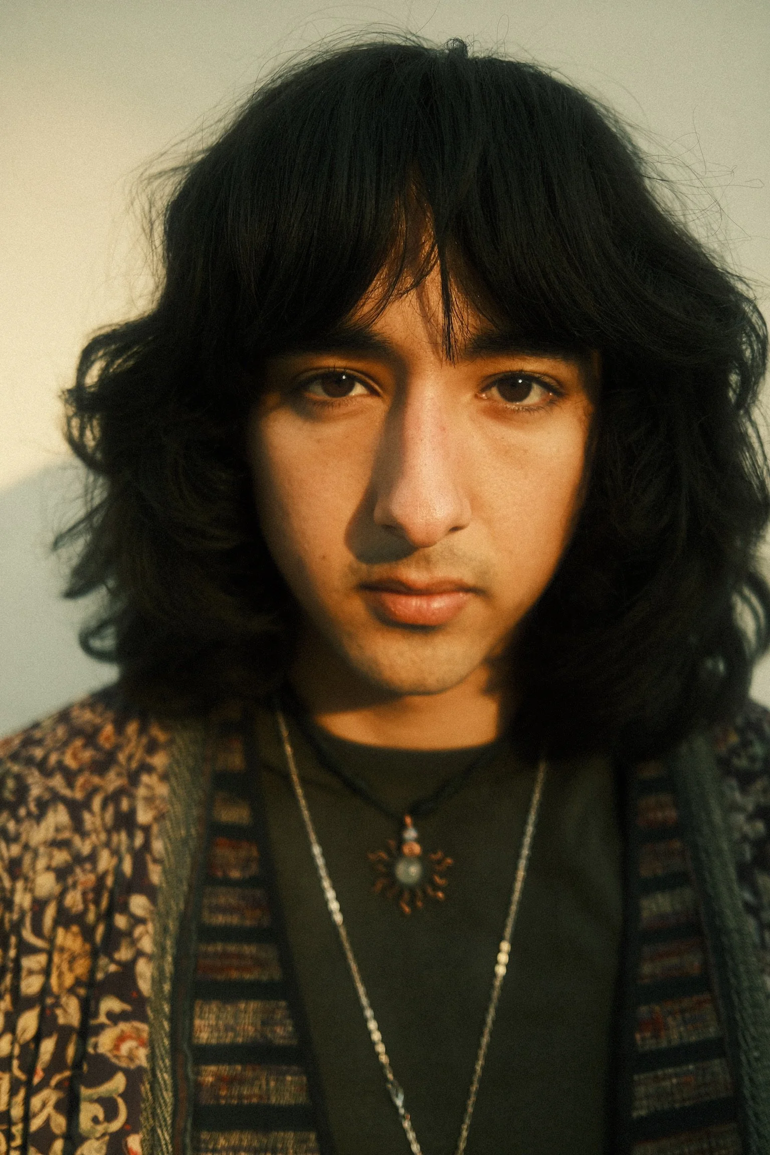 A person with long wavy black hair, brown eyes, wearing a black top, a patterned jacket, and layered necklaces, including a turtle-shaped pendant, against a plain background.