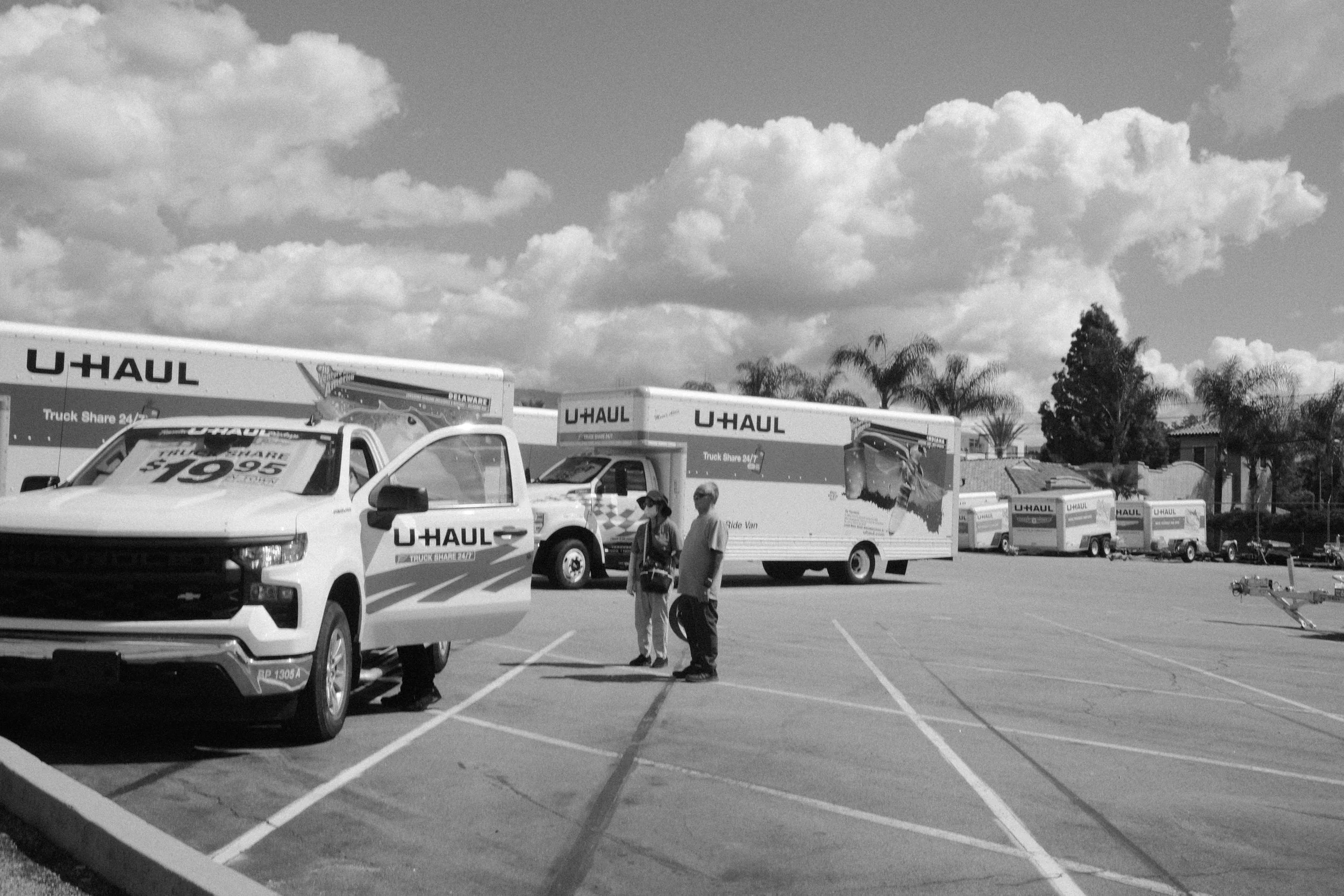Parking lot with U-Haul trucks and vehicles, including a pickup truck and a box truck, under partly cloudy sky, with a few people walking among the vehicles.