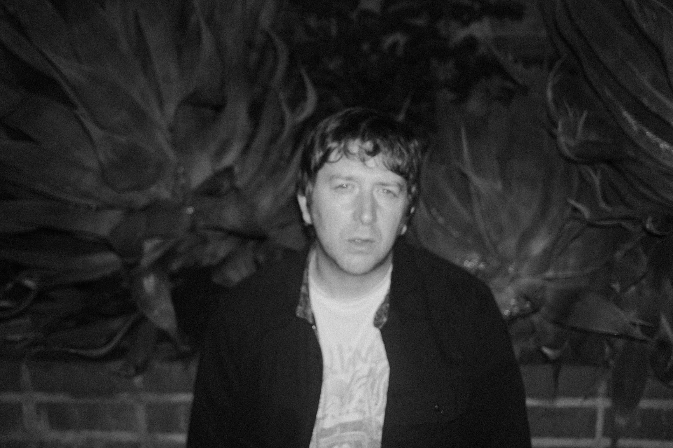 A black and white photo of a young man with curly hair and an intense expression, surrounded by dark leaves or foliage.