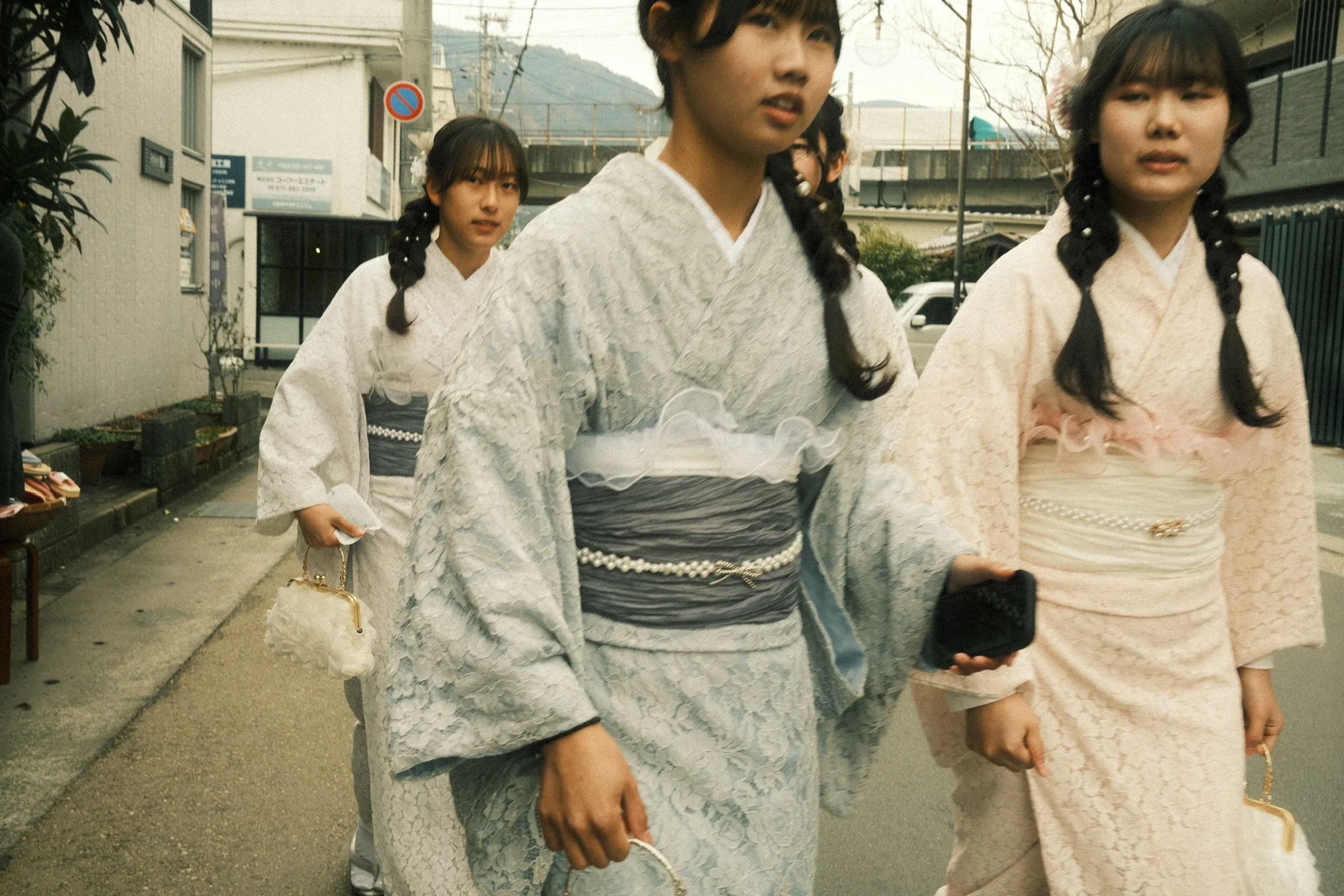 Four women wearing traditional Japanese kimonos walking outdoors on a street with buildings, trees, and mountains in the background.