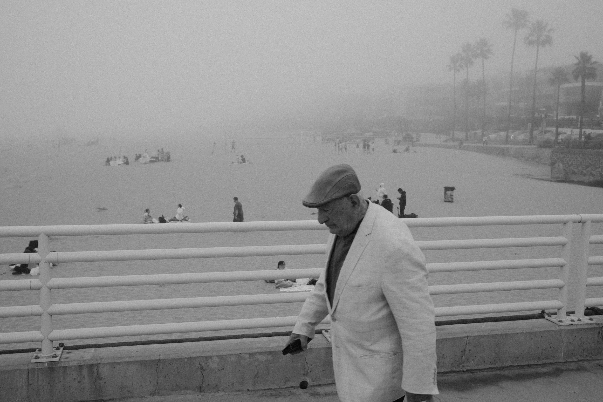 A man in a light-colored suit and cap walking past a white railing on a beach promenade. In the background, people are sitting and walking on the sandy beach, with palm trees and buildings along the coastline. The scene appears hazy or foggy.