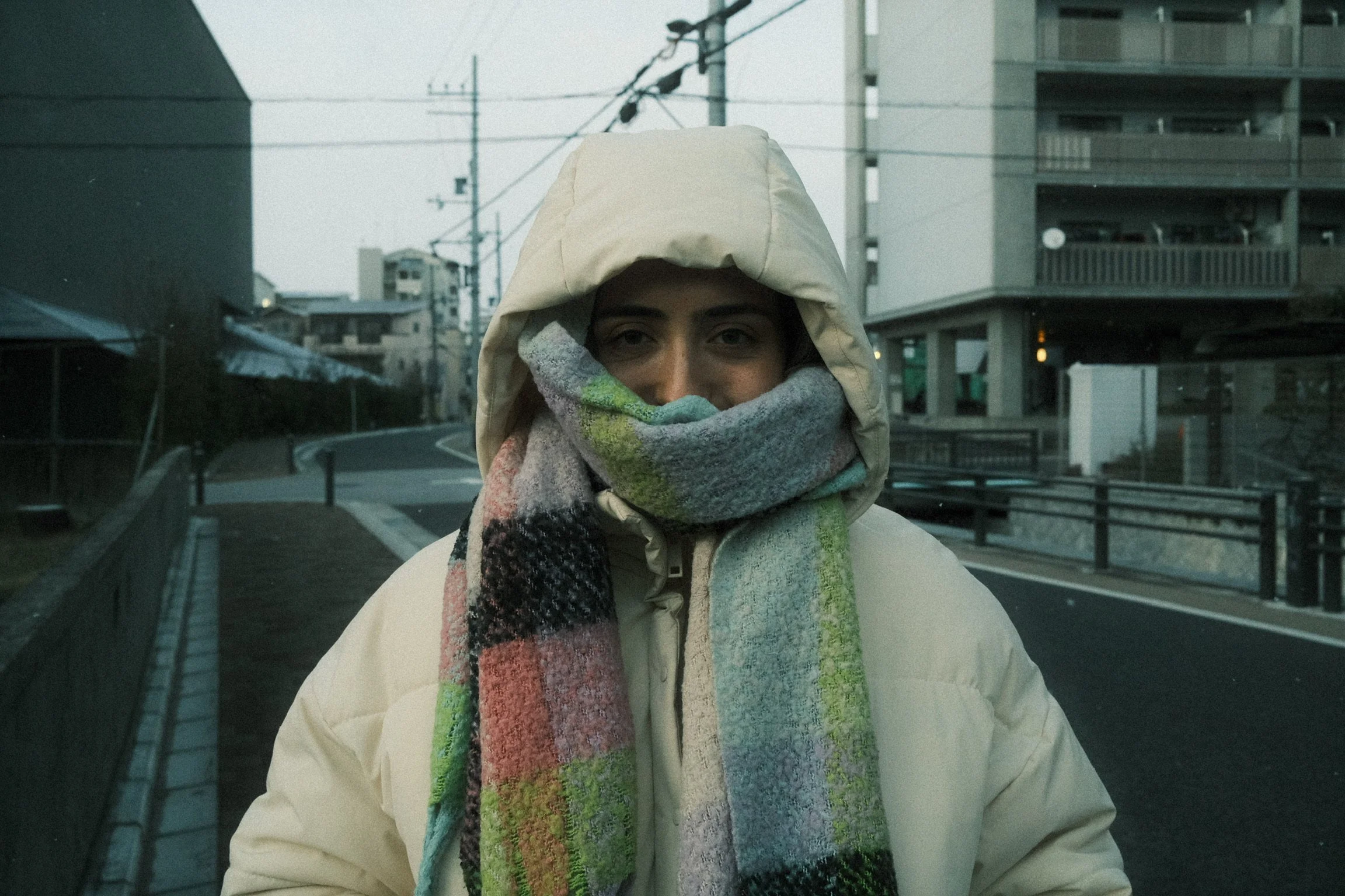 A person wearing a beige hooded jacket and a multicolored scarf covering their mouth and nose standing outdoors on a city street with residential buildings and power lines in the background.