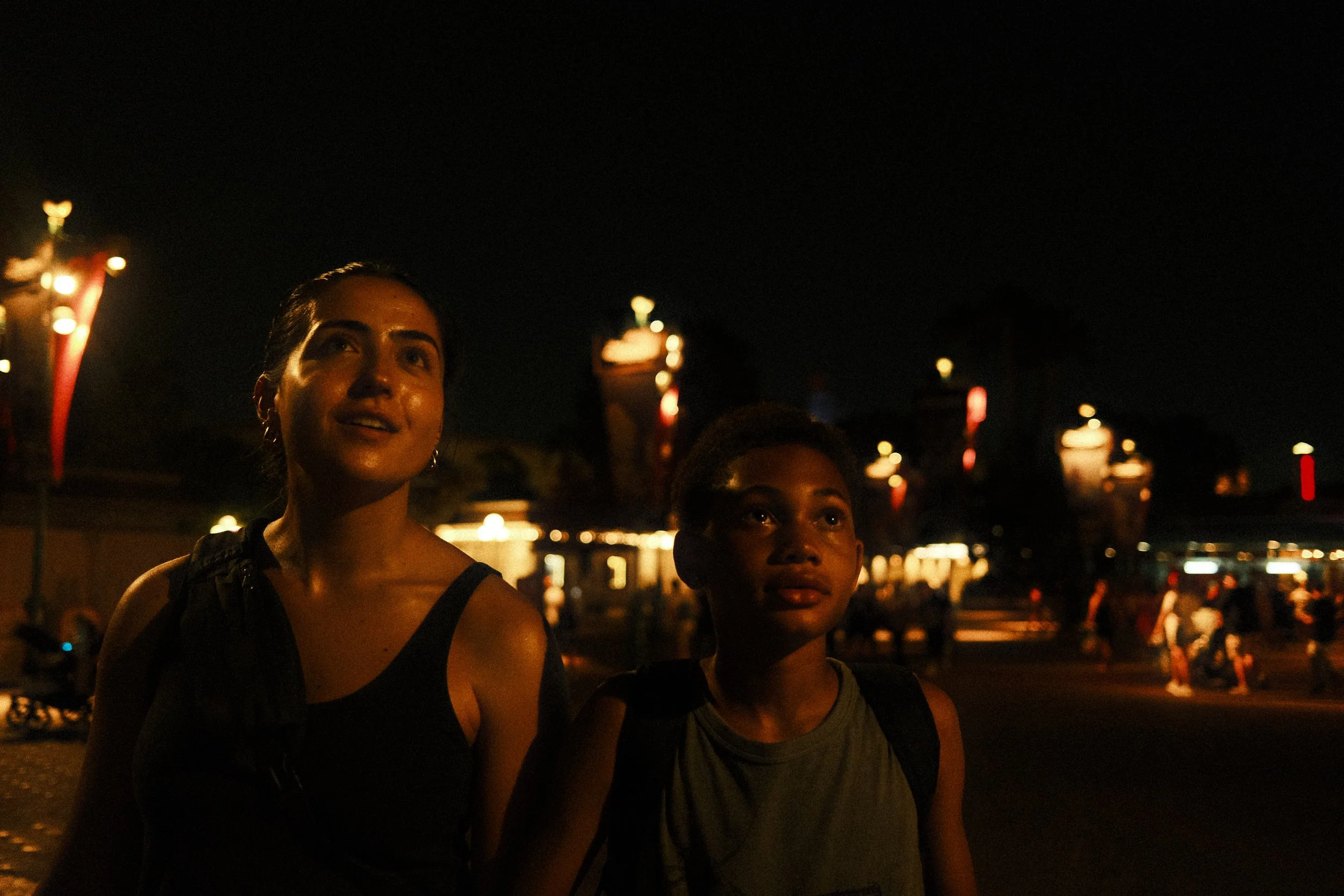 Nighttime scene with three women standing outdoors, illuminated by artificial lights, with a dark sky and brightly lit structures in the background.