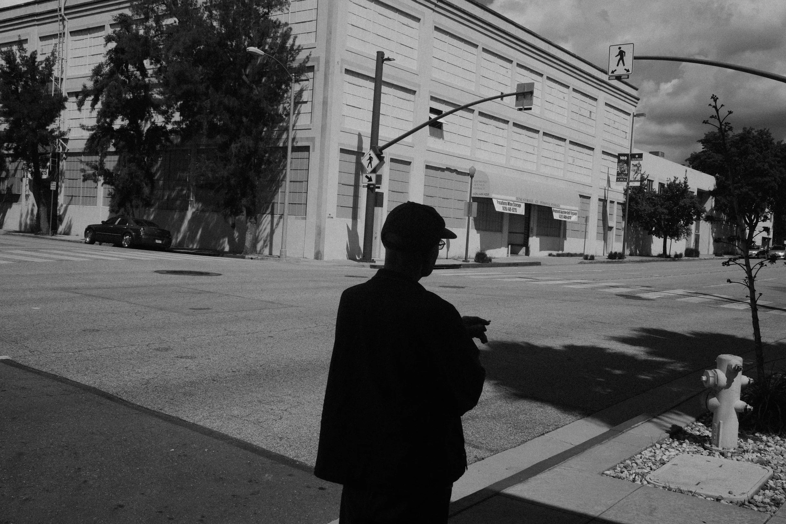 A person wearing a cap and glasses sitting on a sidewalk, with trees and a large building in the background, captured in black and white.