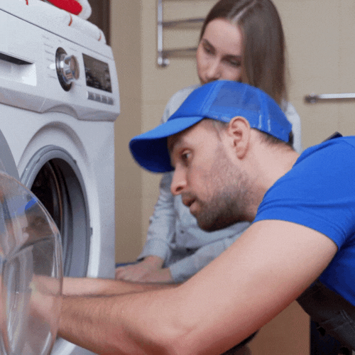 Un hombre con gorra azul arreglando una lavadora con una mujer observando.