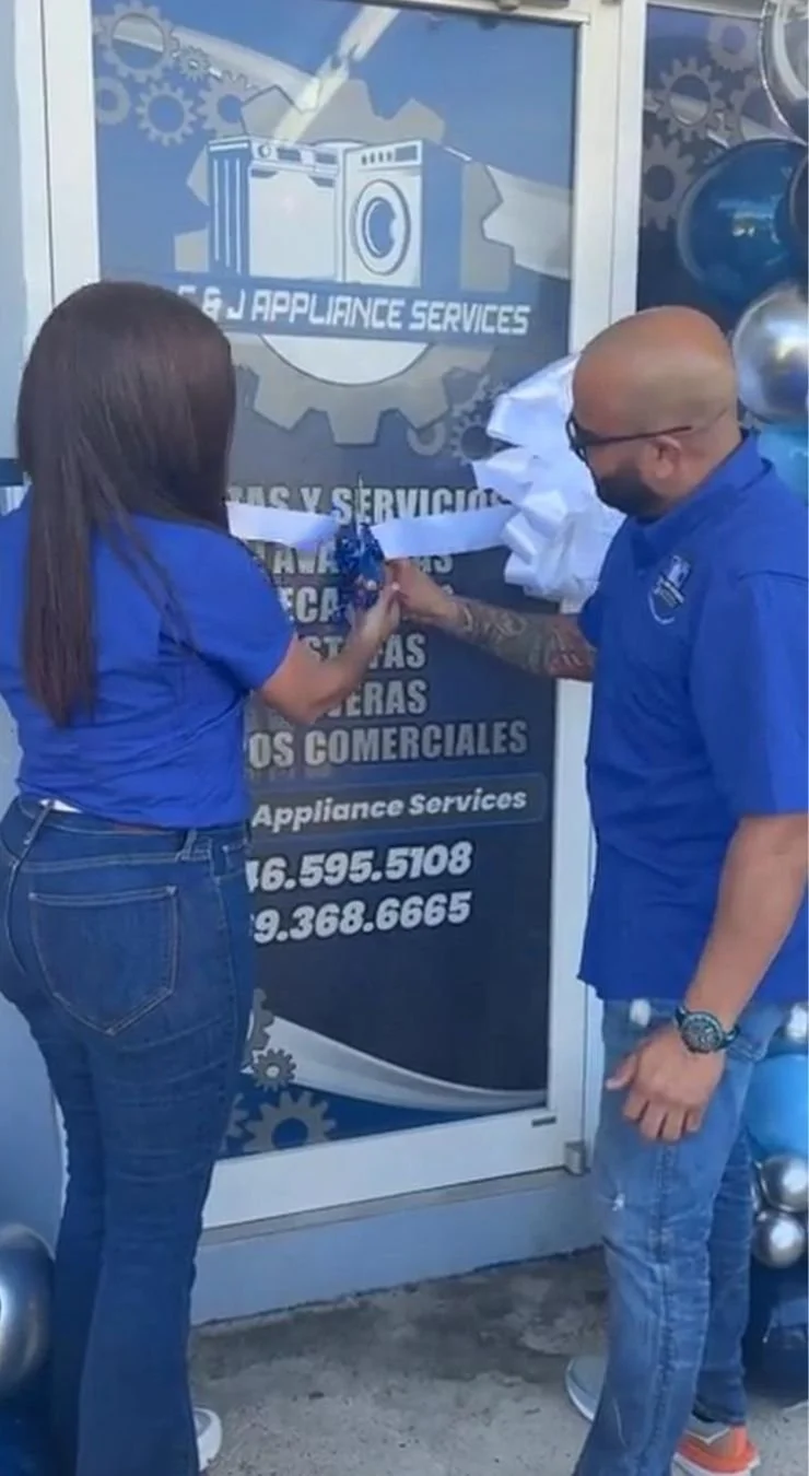 Christian Ortiz y Wilmarie Ortiz, dueños  en uniforme azul realizan la ceremonia de inauguración de C&J Appliance Services un negocio de servicios de electrodomésticos, con globos plateados y un logotipo que muestra una lavadora en el fondo.