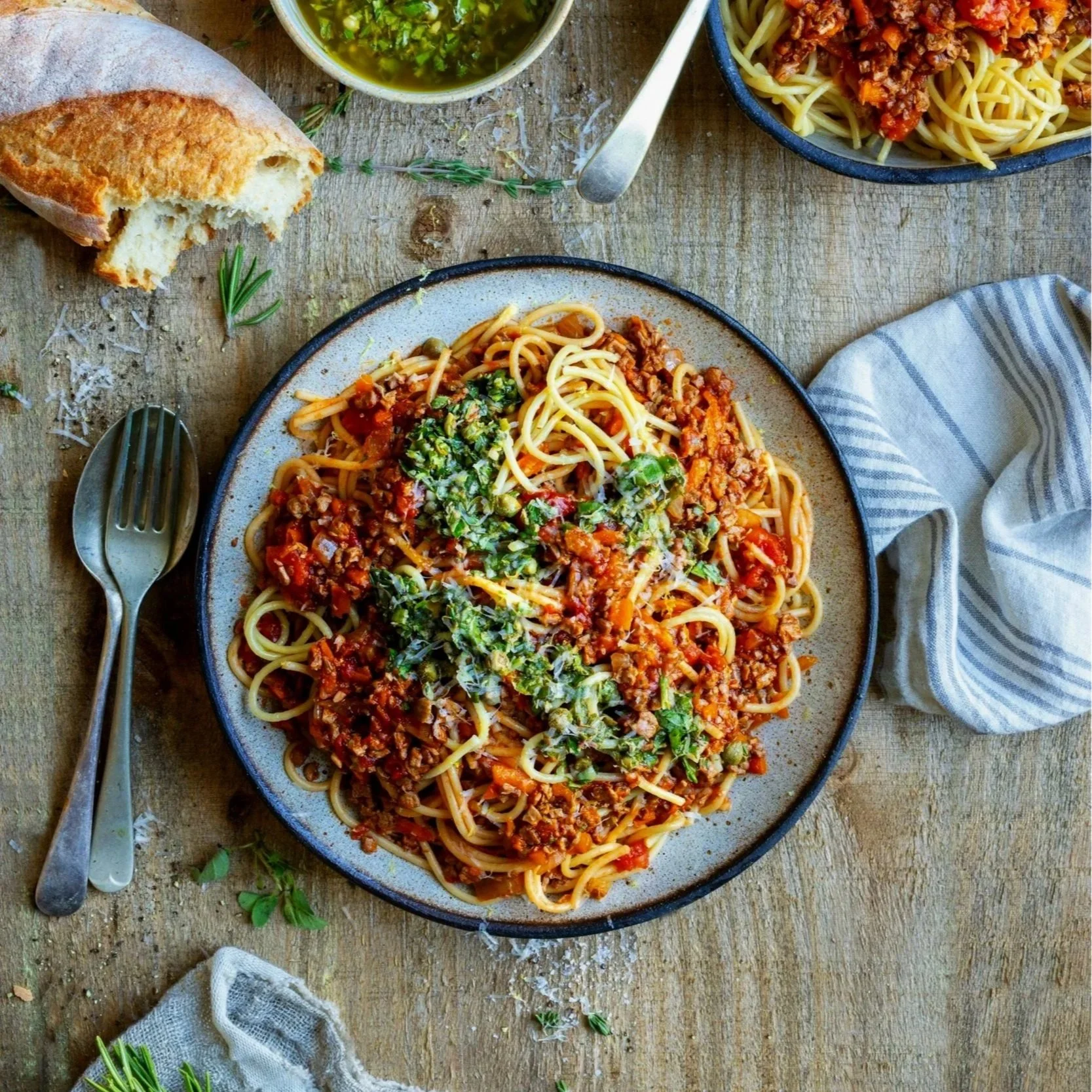 Plate of spaghetti with meat sauce, topped with chopped herbs, with a piece of bread, a fork, a spoon, and a side of green herb sauce on a rustic wooden table.