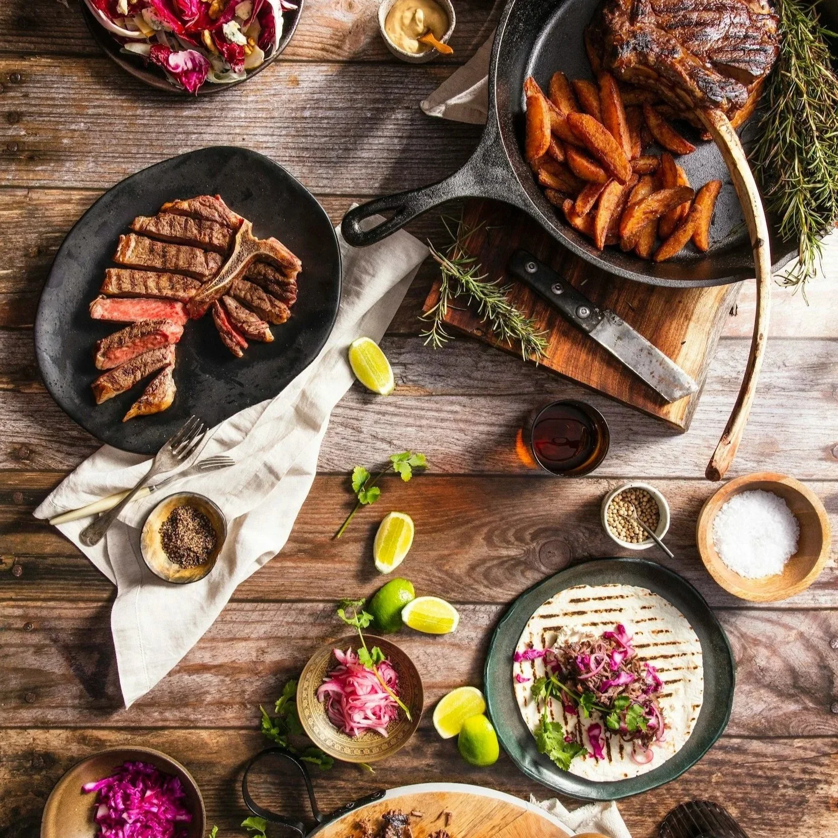 A rustic wooden table set with grilled ribeye steak, roasted potato wedges, fresh lime wedges, a bowl of grilled meat on flatbread with herbs, a salad with purple cabbage, pickled onions, and cilantro, a small bowl of salt, and various seasonings, with drinks and garnishes.