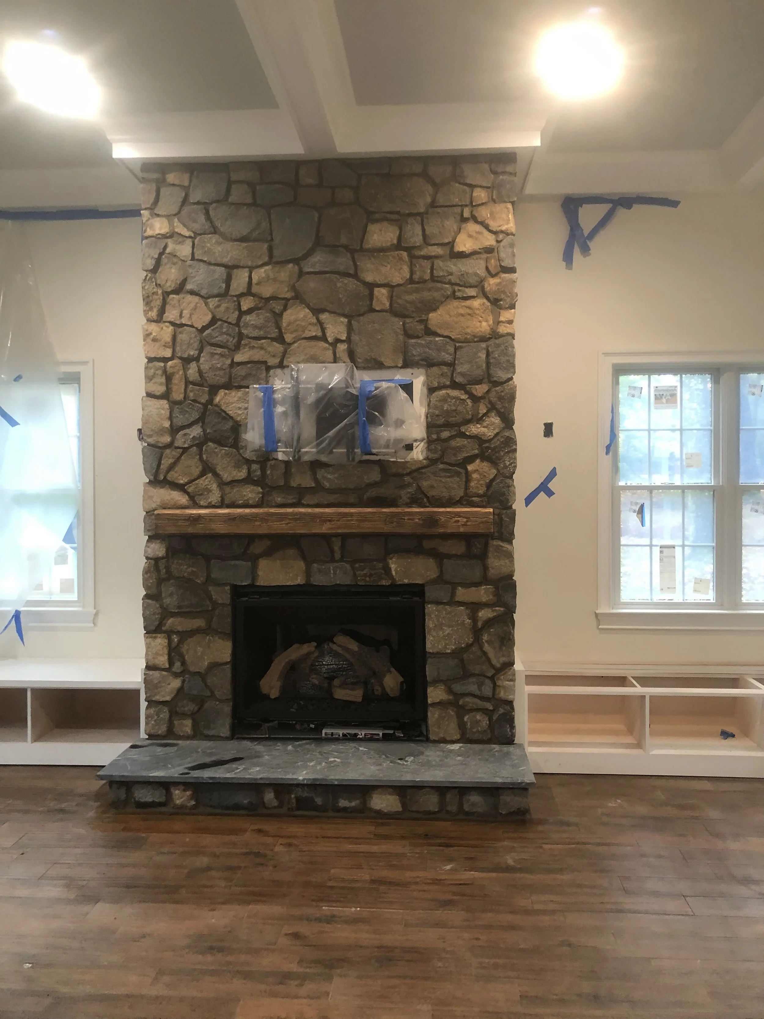 A large stone fireplace in the center of a living room, with a wooden mantle and a stone hearth on a wooden floor. Two windows on either side are covered with painter's tape, indicating ongoing renovation or painting.