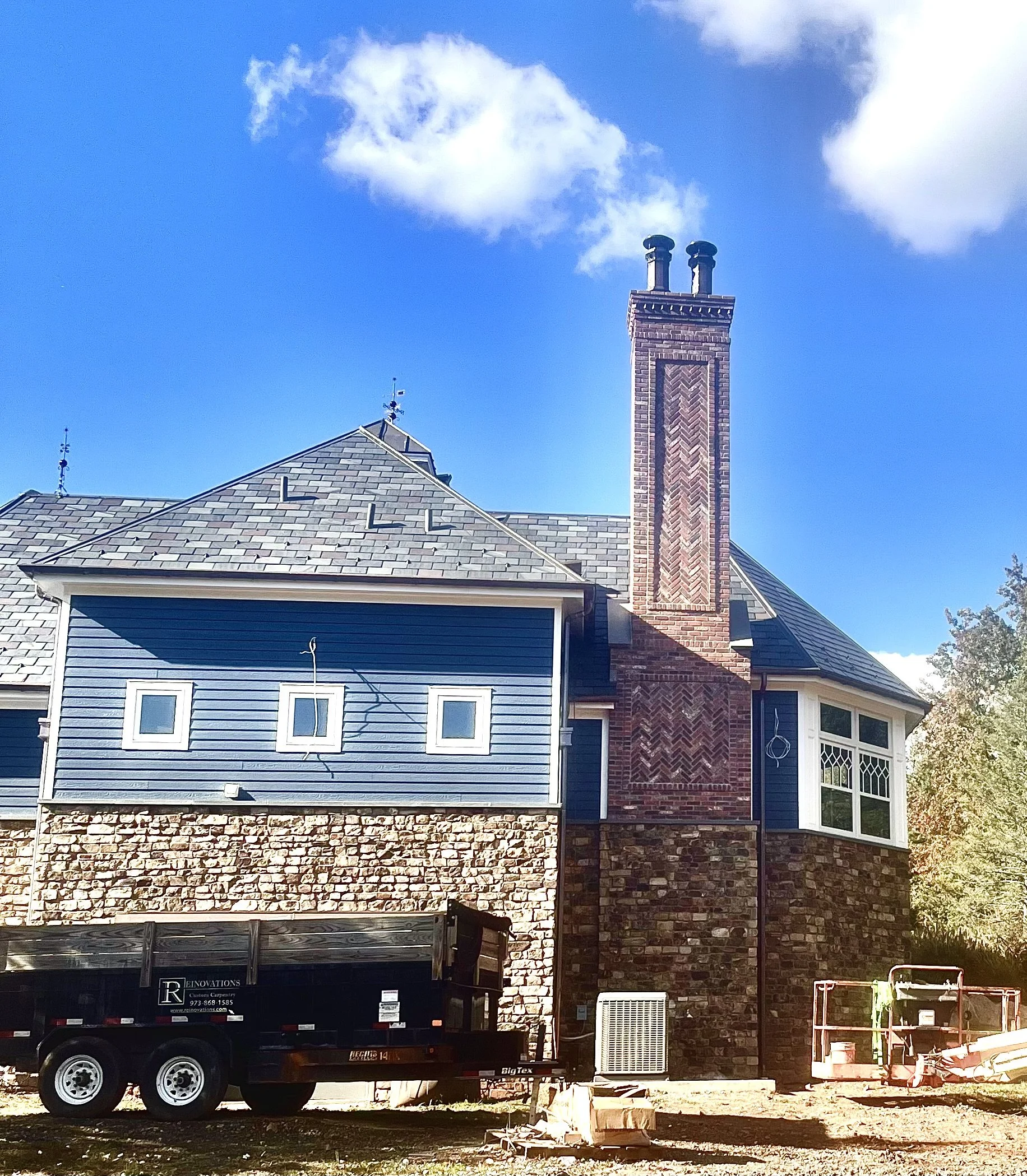 The image shows the back of a house with a combination of brick and blue siding. The house has multiple chimney stacks on the roof, a pitched roof with gray shingles, and a large windowed sunroom on the right. In the foreground, there is a black util