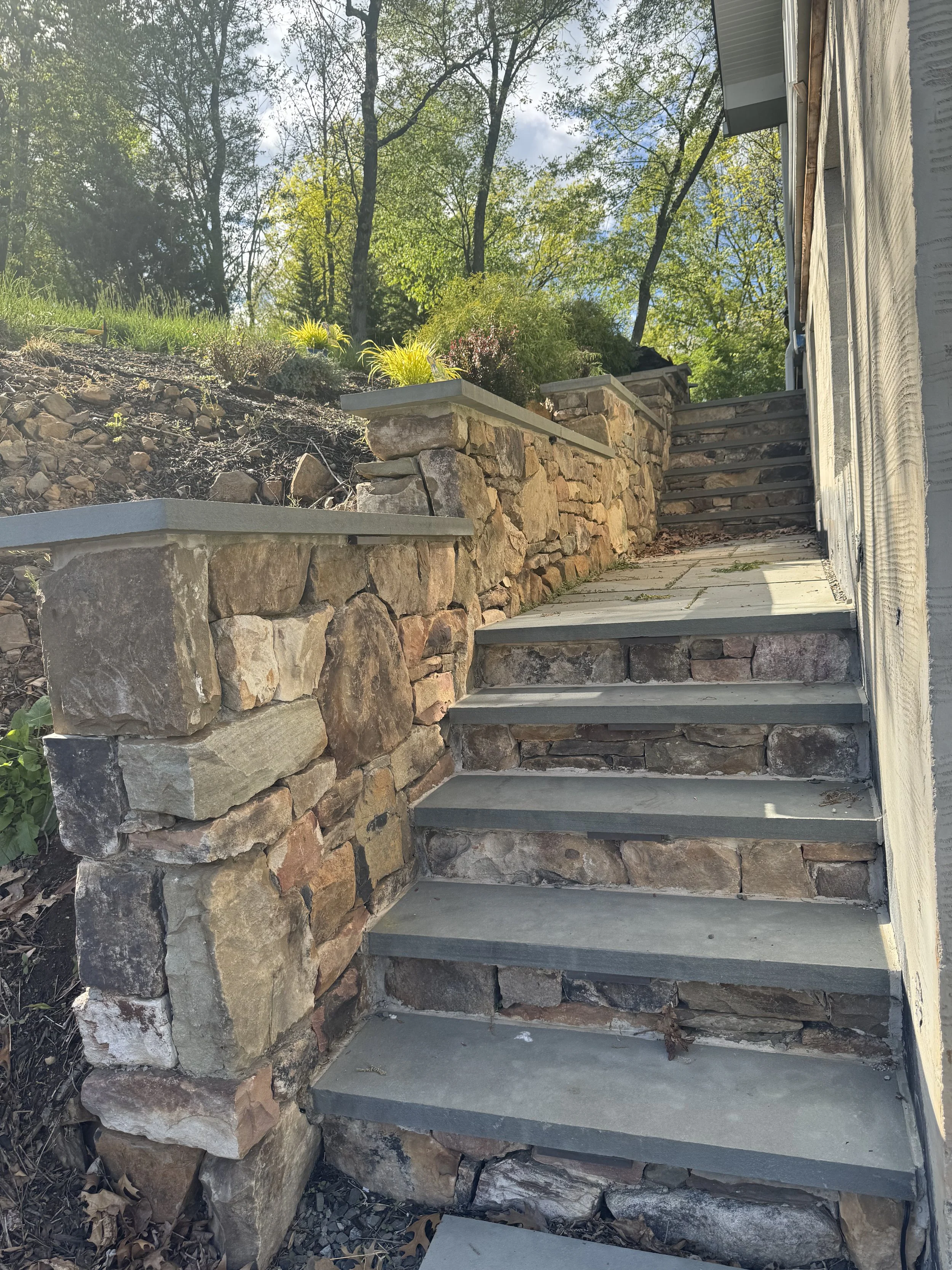 Stone retaining wall with concrete stairs leading upward, surrounded by trees and greenery outdoors.
