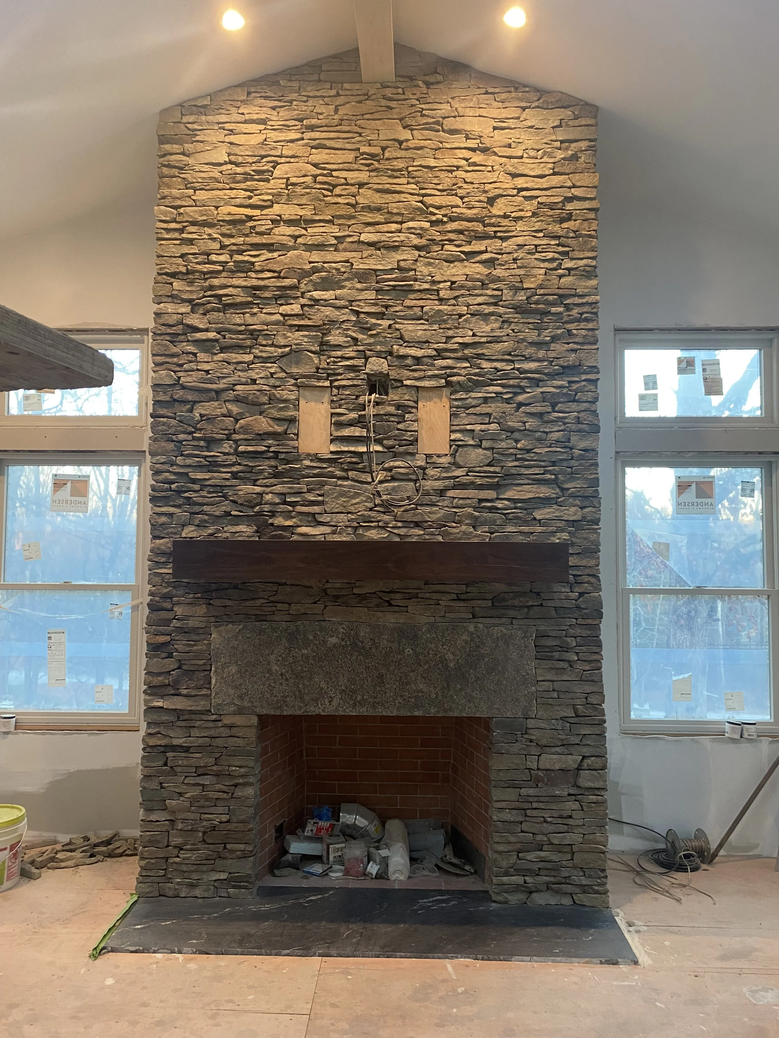 A stone fireplace under construction with an unfinished interior and construction tools in the firebox, in a room with windows and drywall installed, but with tools and materials on the floor.