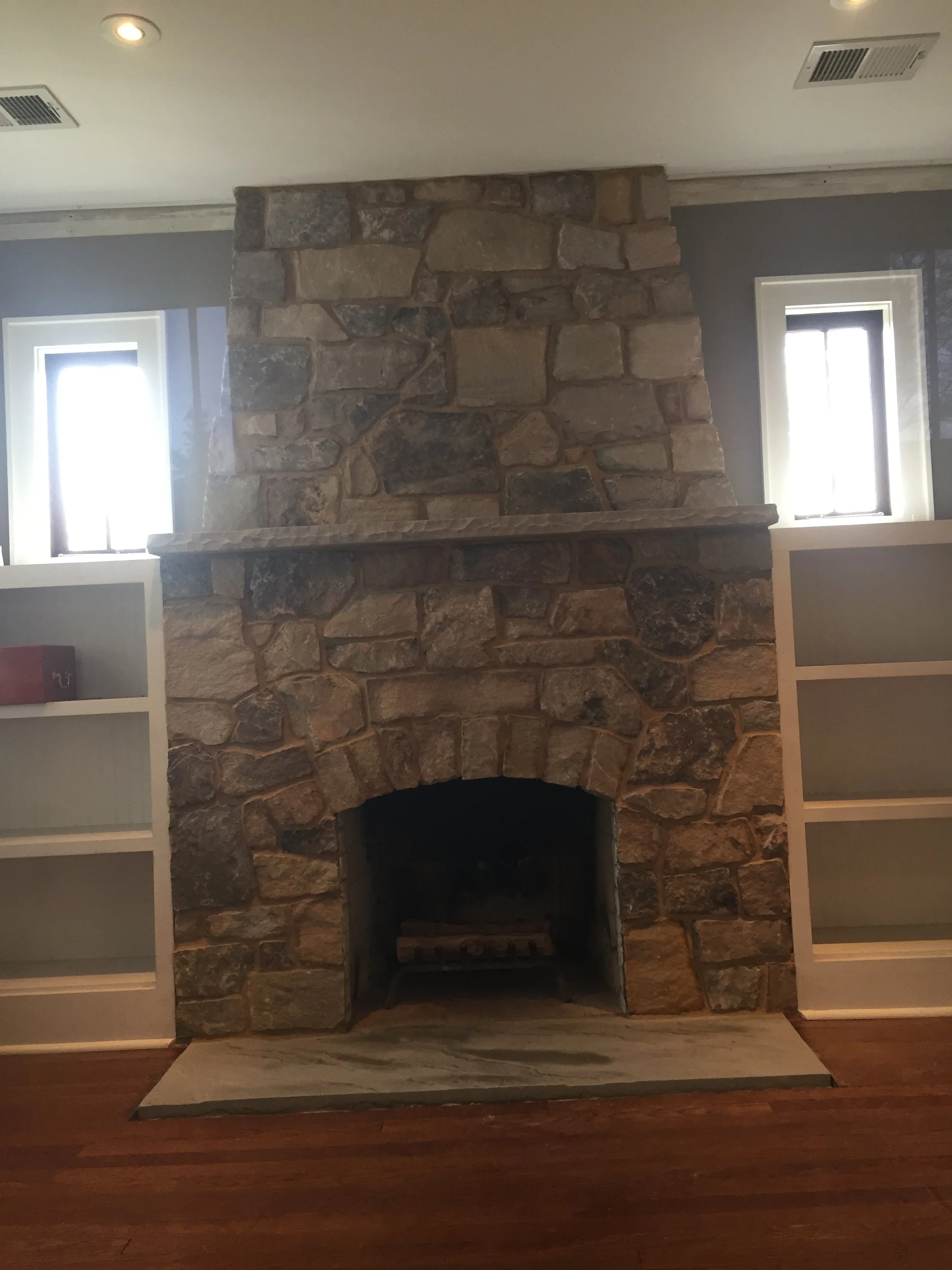 Interior of a room with a large stone fireplace, two tall windows on either side, white built-in shelves, and wooden flooring.