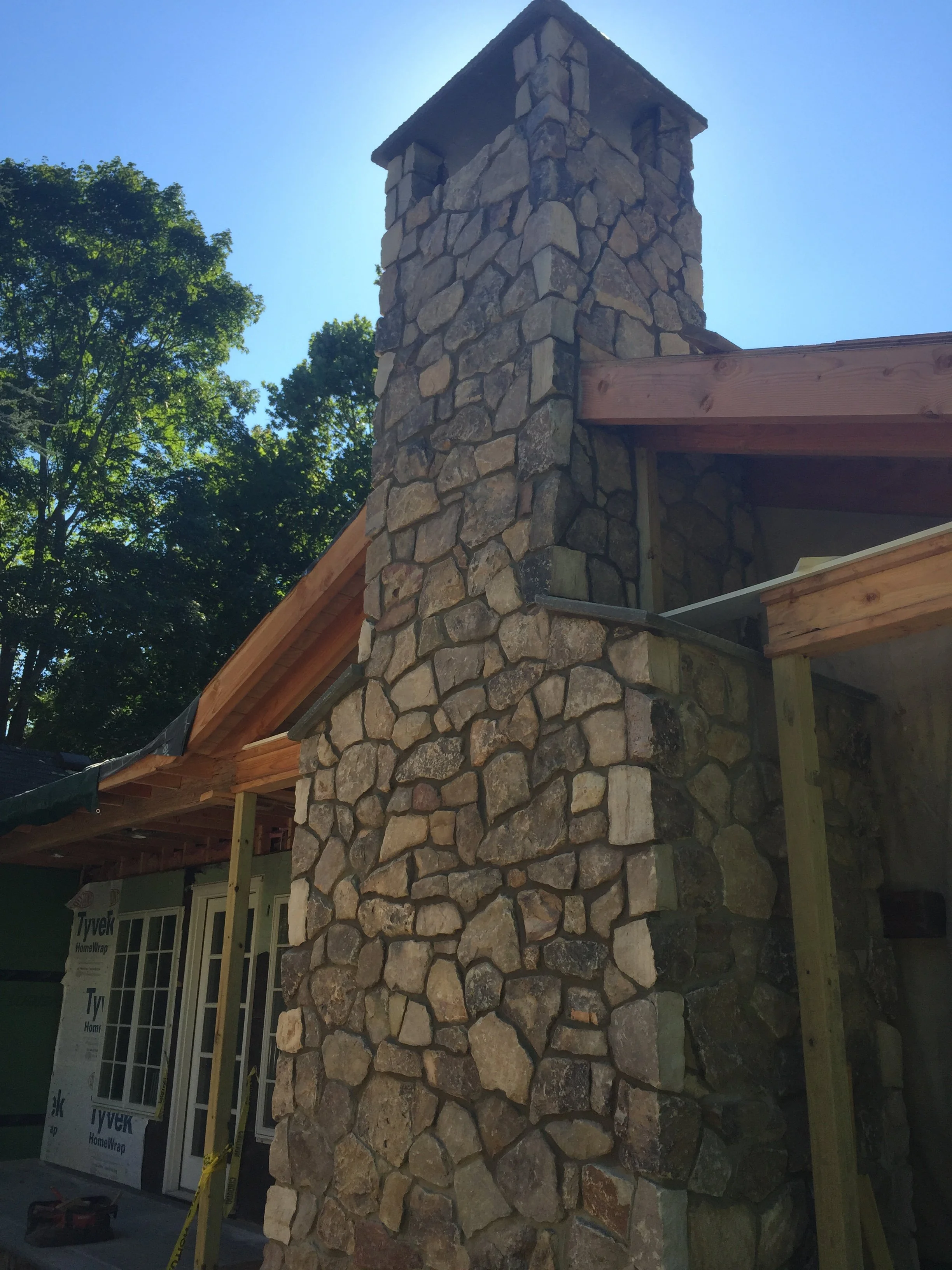 A stone chimney on a house under construction, with a clear blue sky and trees in the background.