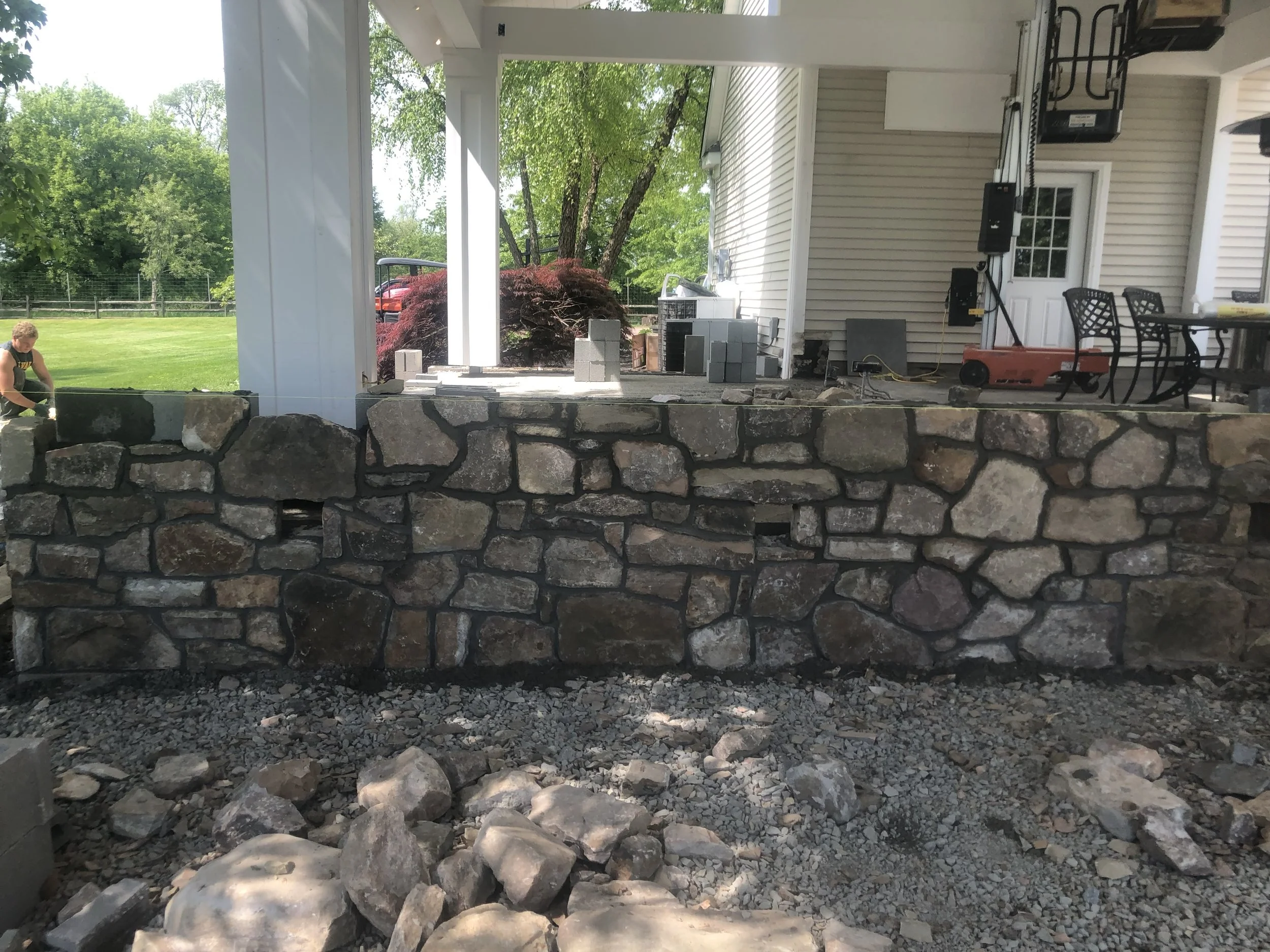Back patio under construction, with stacked stones forming a low wall, and construction materials, an orange air compressor, and a table with black chairs.