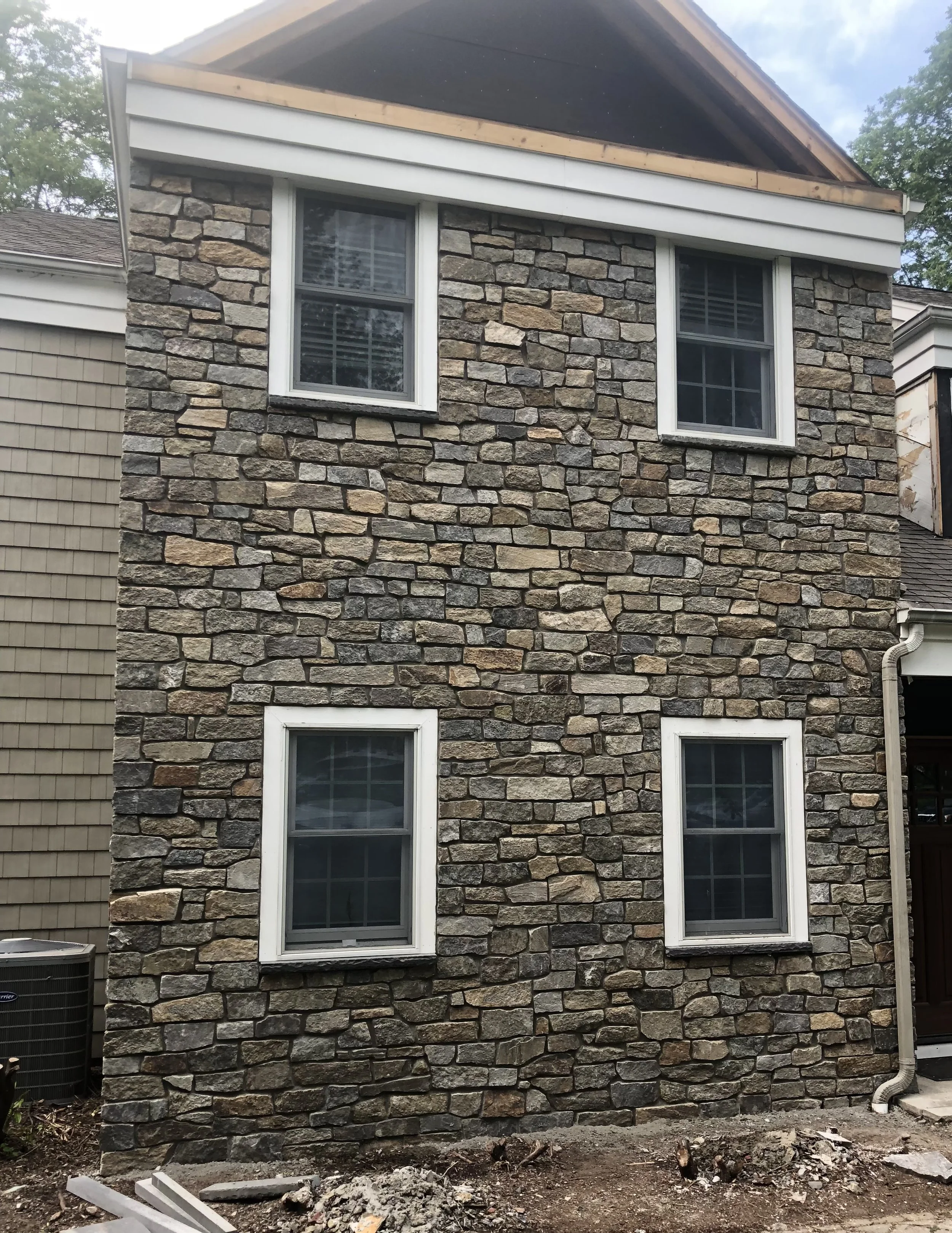 Back of a house with stone siding, two windows with white trim, and partial construction work on the roof. There is dirt and construction debris on the ground.