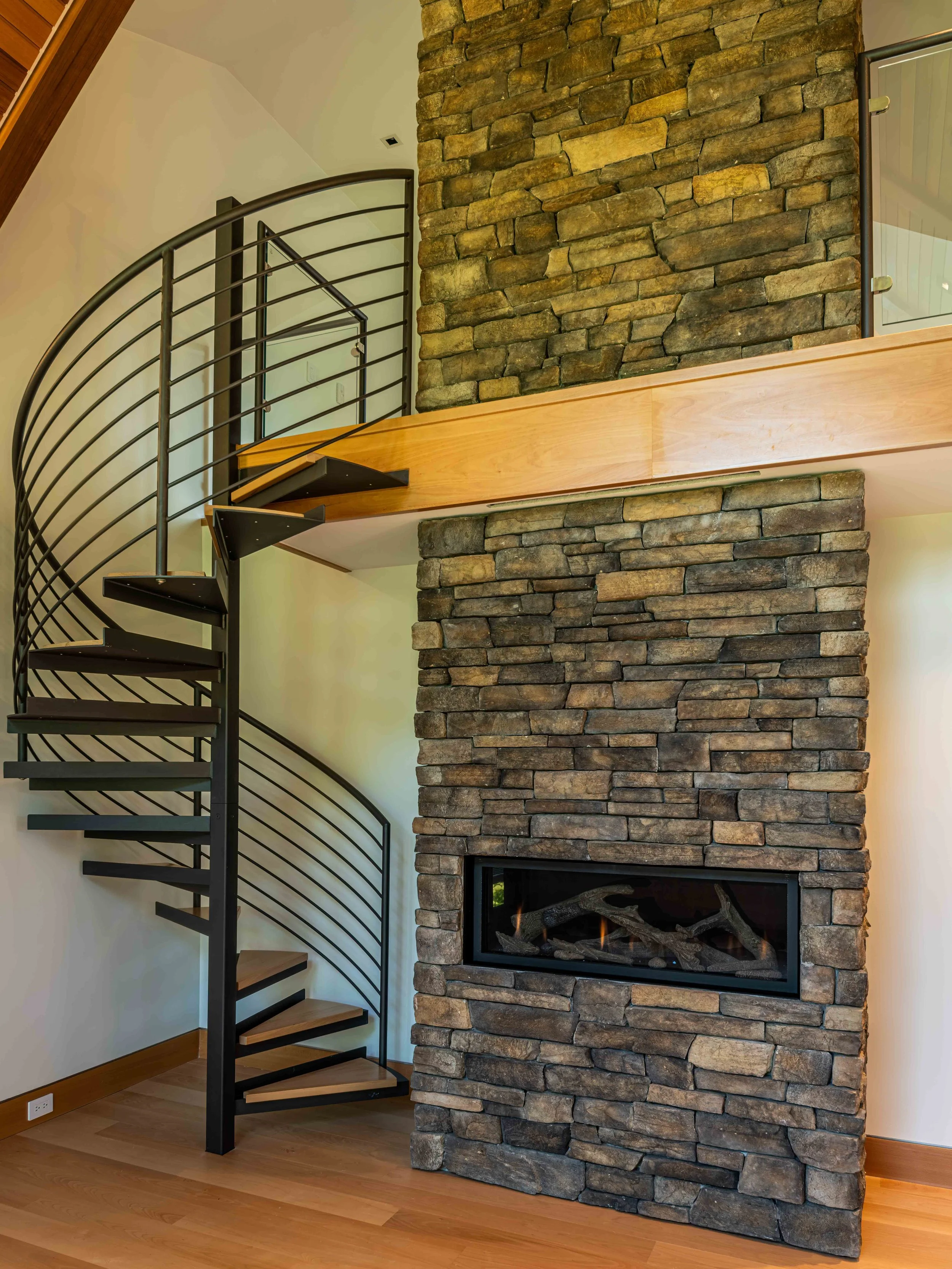 Interior of a room featuring a spiral staircase with wooden steps and black metal railing, a stone fireplace withfire logs, and hardwood flooring.