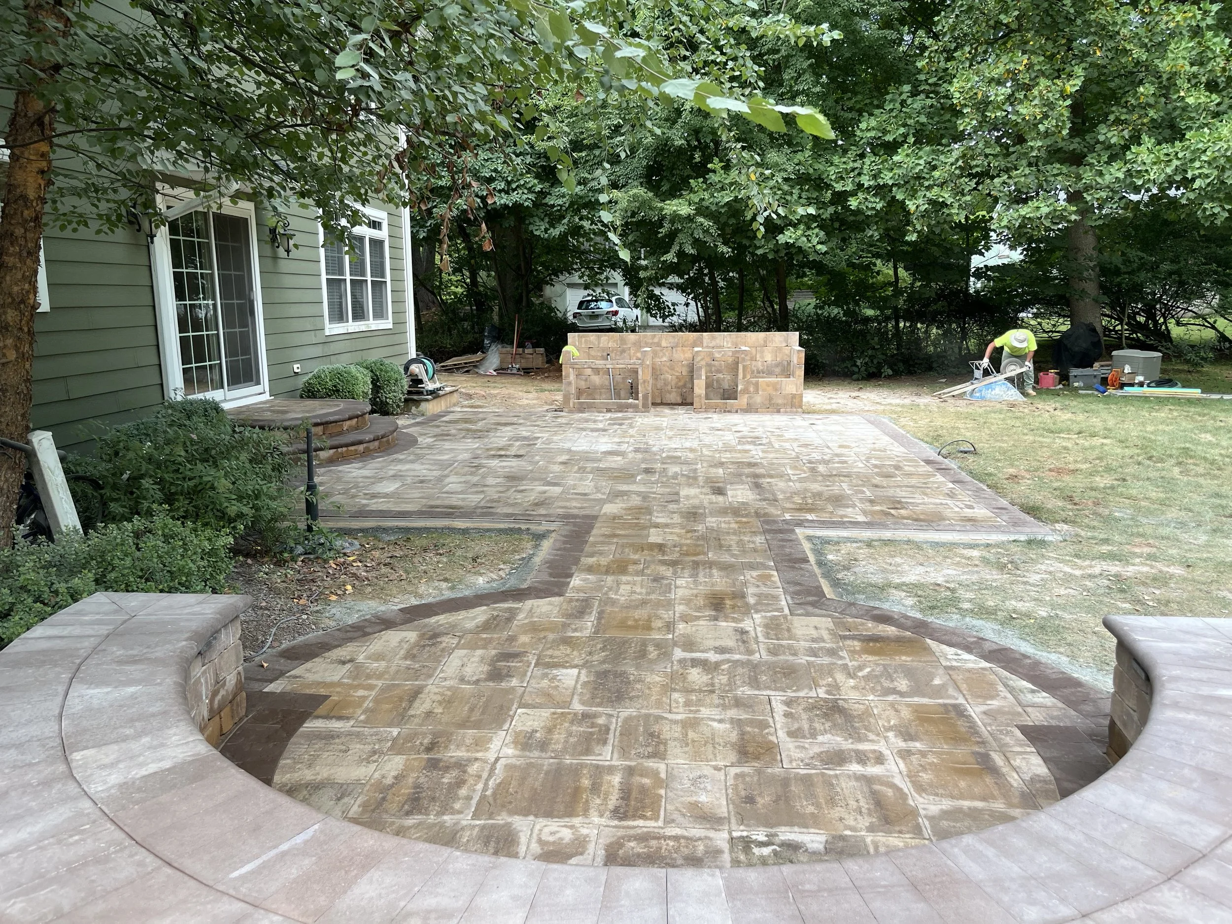 Backyard with newly installed brick and stone patio, green house, trees, landscaping tools, and a person working near a storage container.
