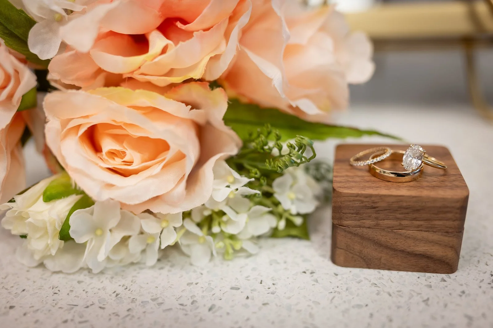 A bouquet of pink roses and white flowers next to two rings, one with a large central diamond and the other with smaller diamonds, on a wooden block.