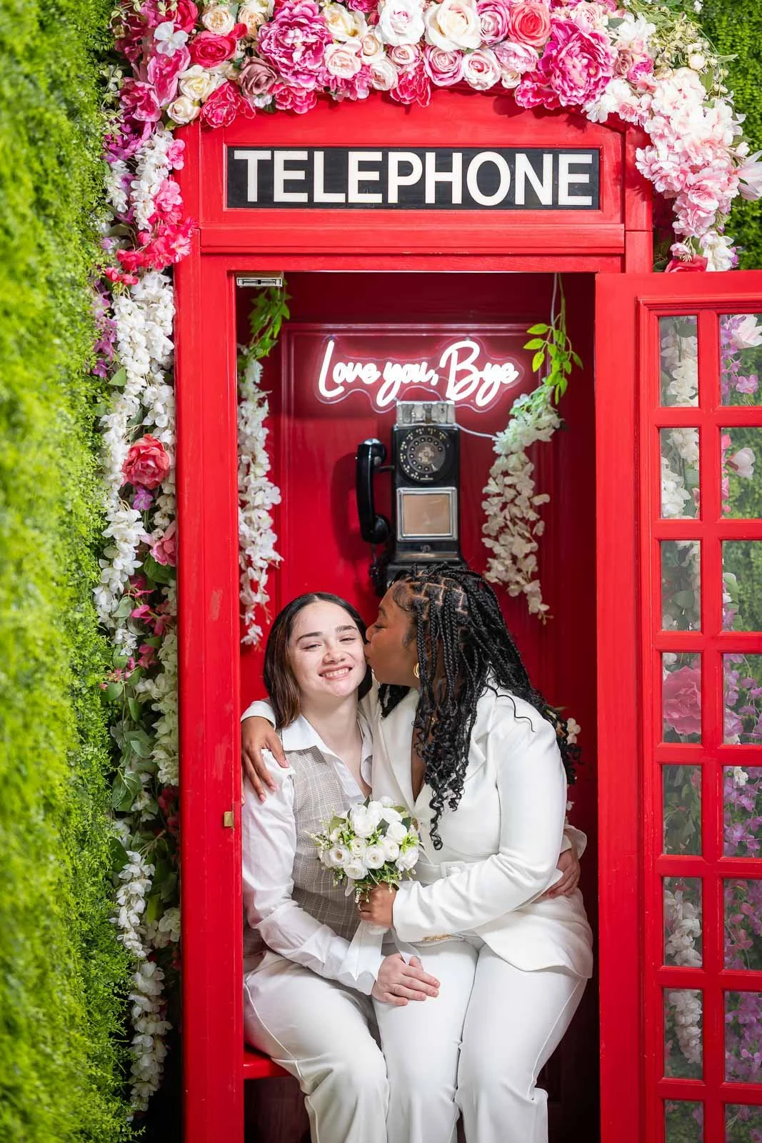 Two women sitting inside a vintage red telephone booth decorated with pink and white flowers, one woman is kissing the other on the cheek, who is holding a bouquet of white roses, with a neon sign saying 'Love you. Bye' inside.