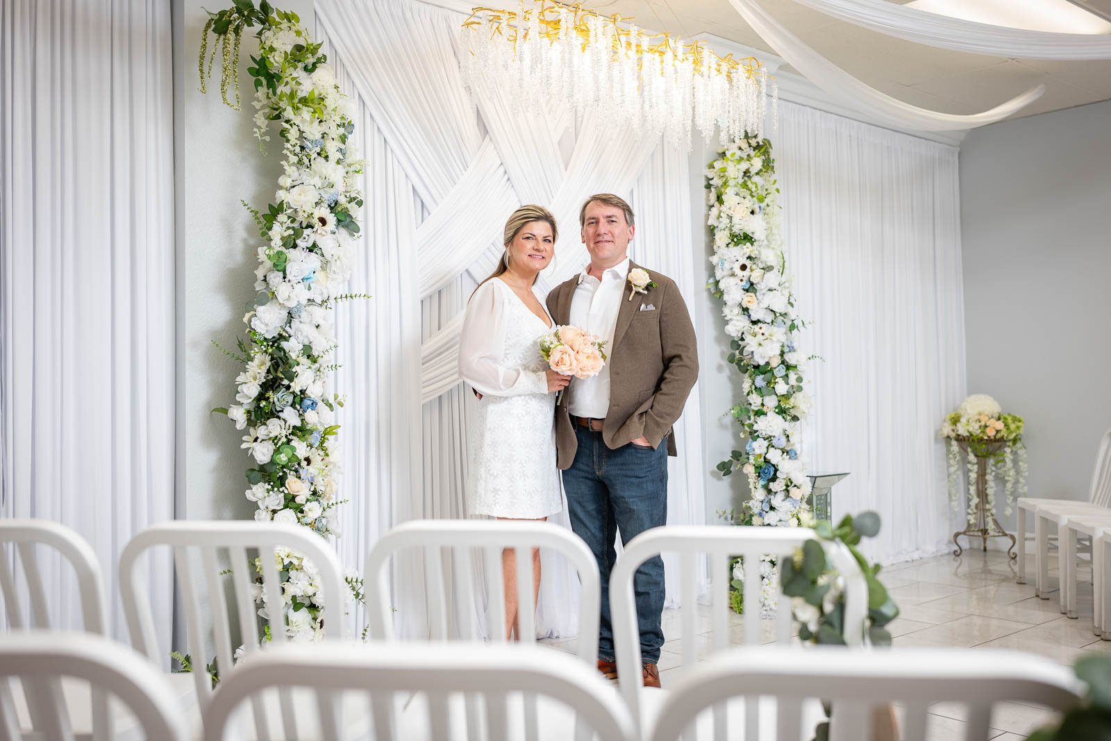A couple standing in front of wedding arch decorated with white flowers and drapes, posing for a picture at a wedding ceremony.
