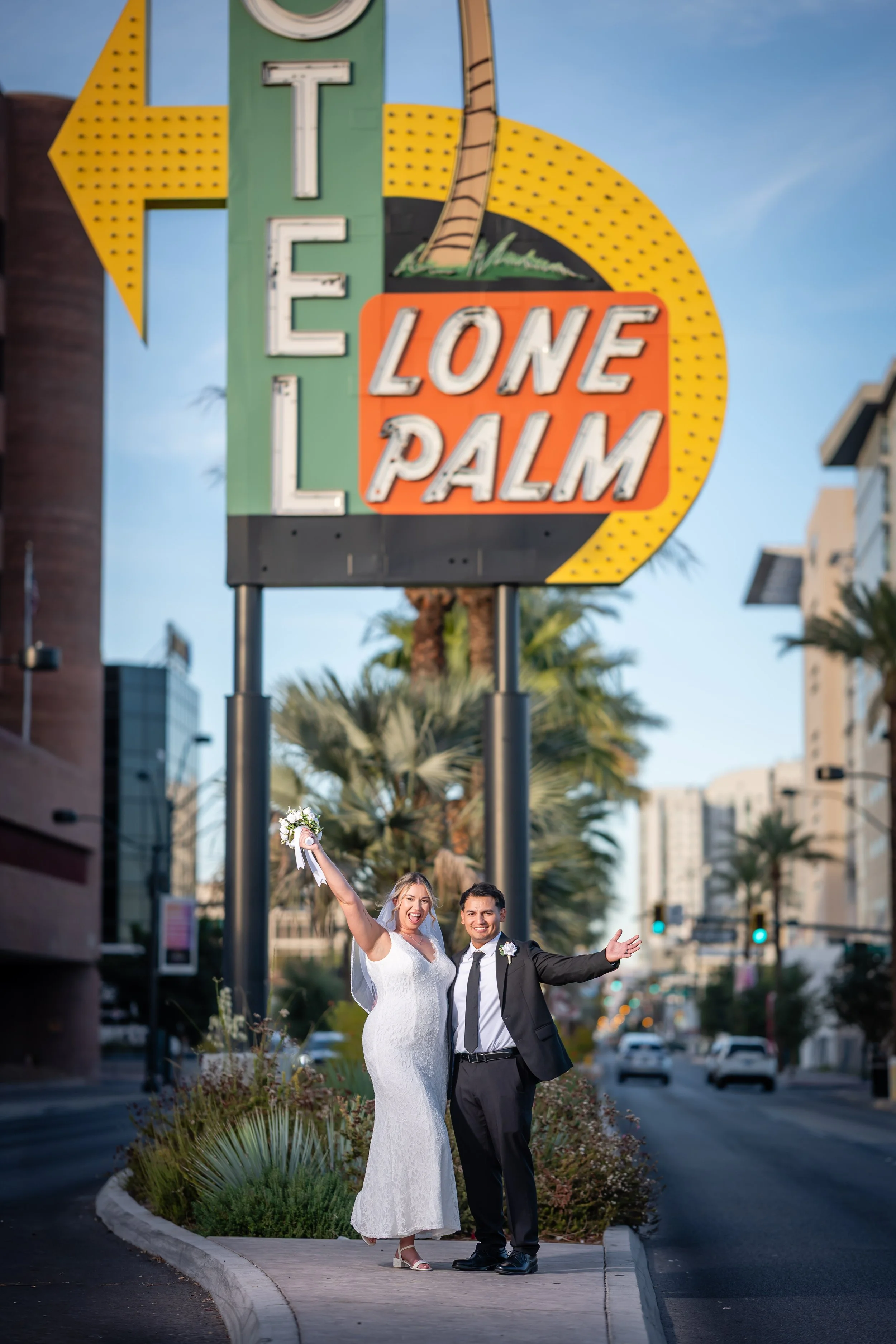 A newlywed couple stands on a sidewalk in front of the Lone Palm sign in Las Vegas, celebrating with their arms raised and smiling.