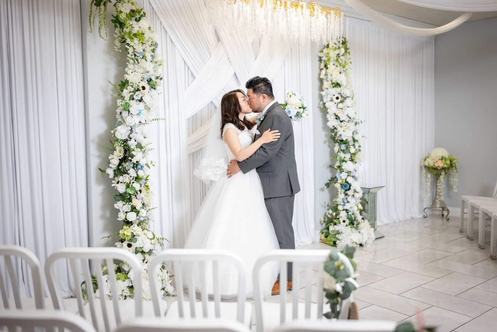 A bride and groom kissing during their wedding ceremony, standing under a decorated white floral arch with white flowers and greenery, in a room with white drapes and tiled floor.