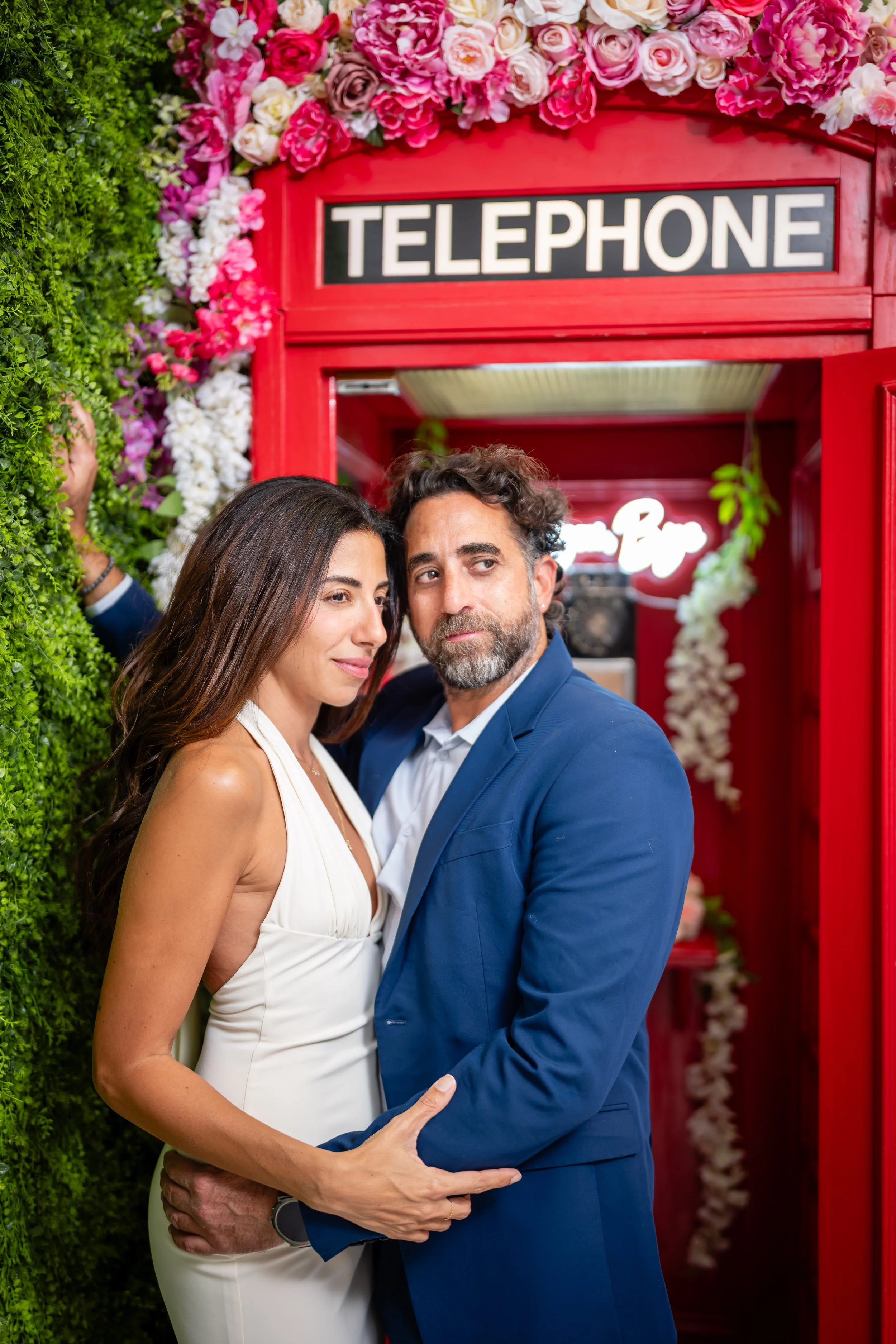 A couple standing close together at a photo booth with a red British-style telephone booth decorated with pink and white flowers behind them.