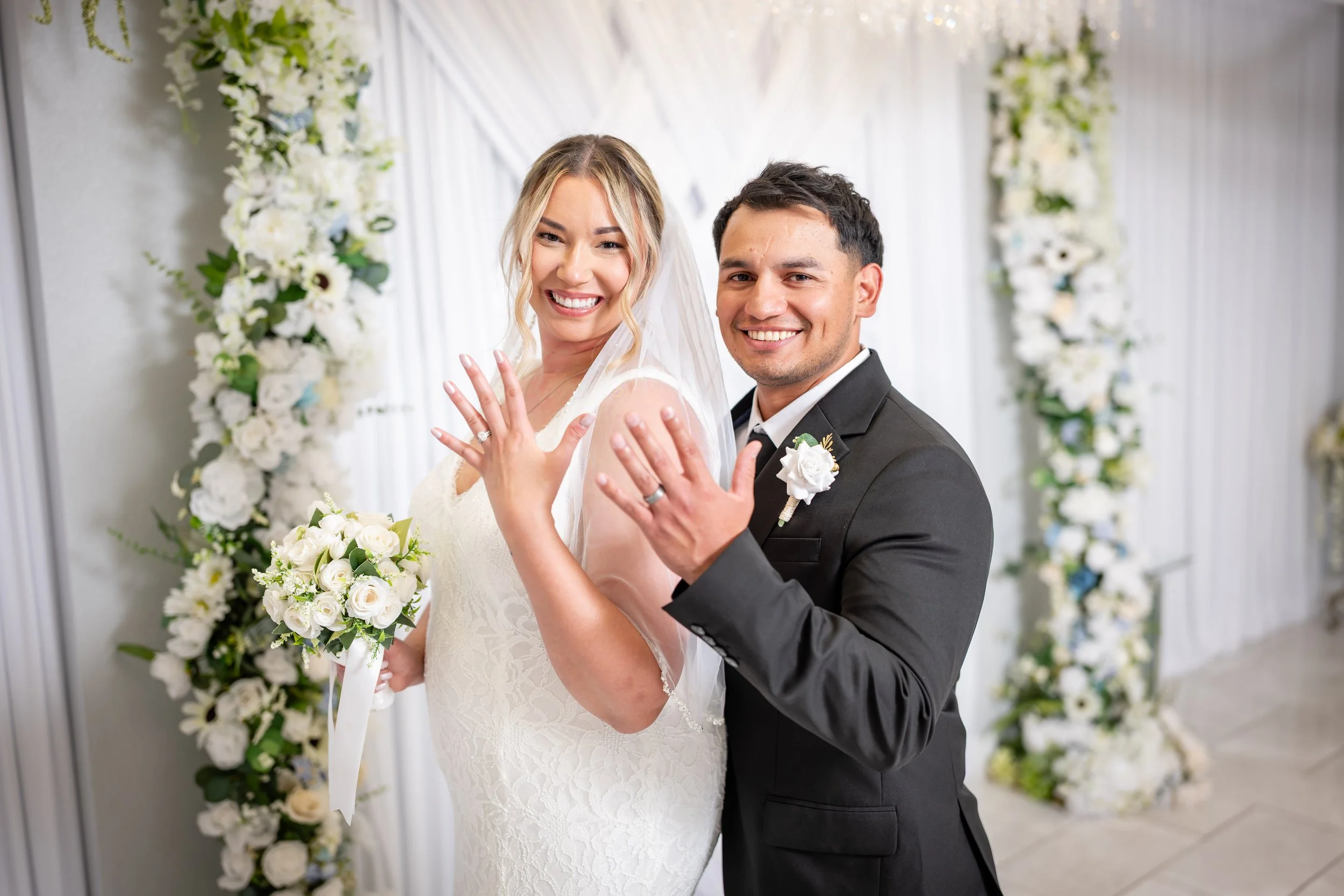 Happy bride and groom showing off their wedding rings in front of floral arch at wedding ceremony