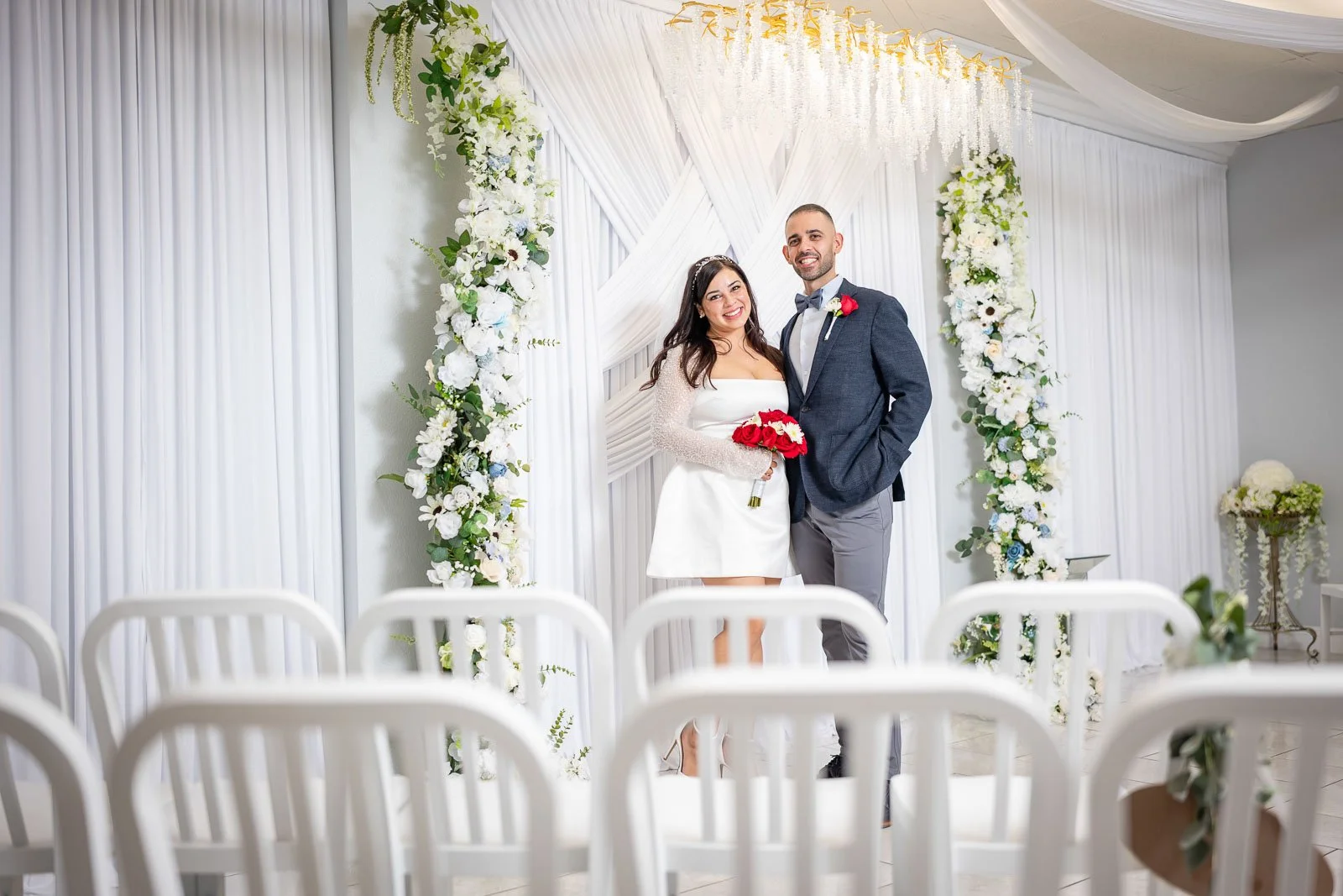 A bride and groom stand together smiling inside a decorated wedding arch, holding a bouquet of red roses, during their wedding ceremony. The background features white draped fabric and floral arrangements.