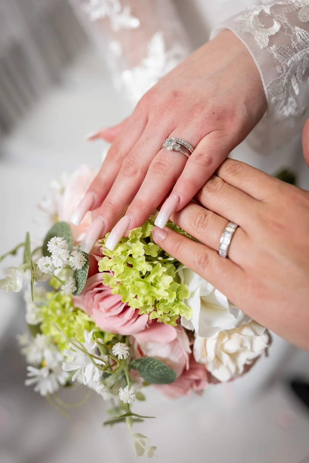 Close-up of a bride and groom's hands showing wedding rings, with a bridal bouquet of pink and white flowers and greenery.