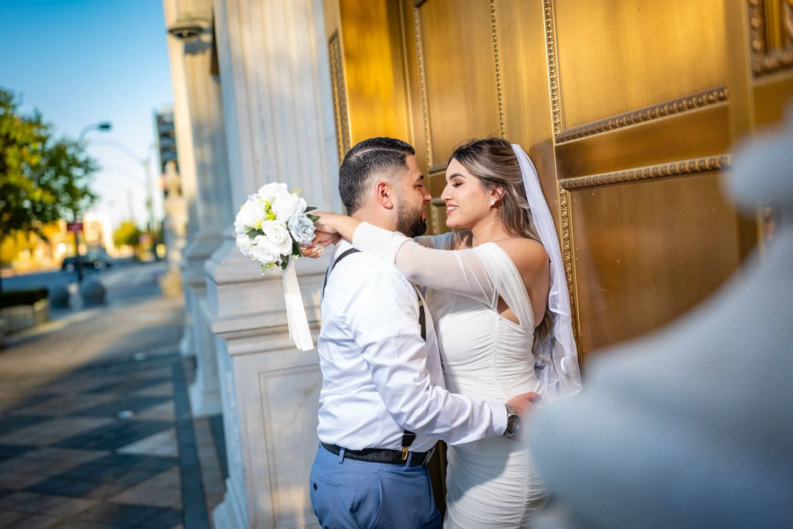 A bride and groom standing close, smiling and touching noses, with the bride holding a bouquet of white flowers, against a golden architectural backdrop outside during sunset.