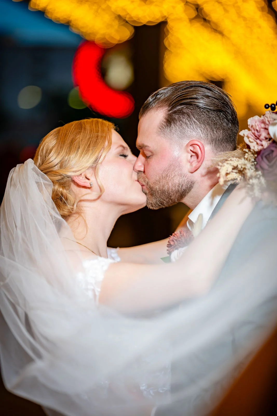 A bride and groom kiss during their wedding reception, with blurred holiday lights in the background.