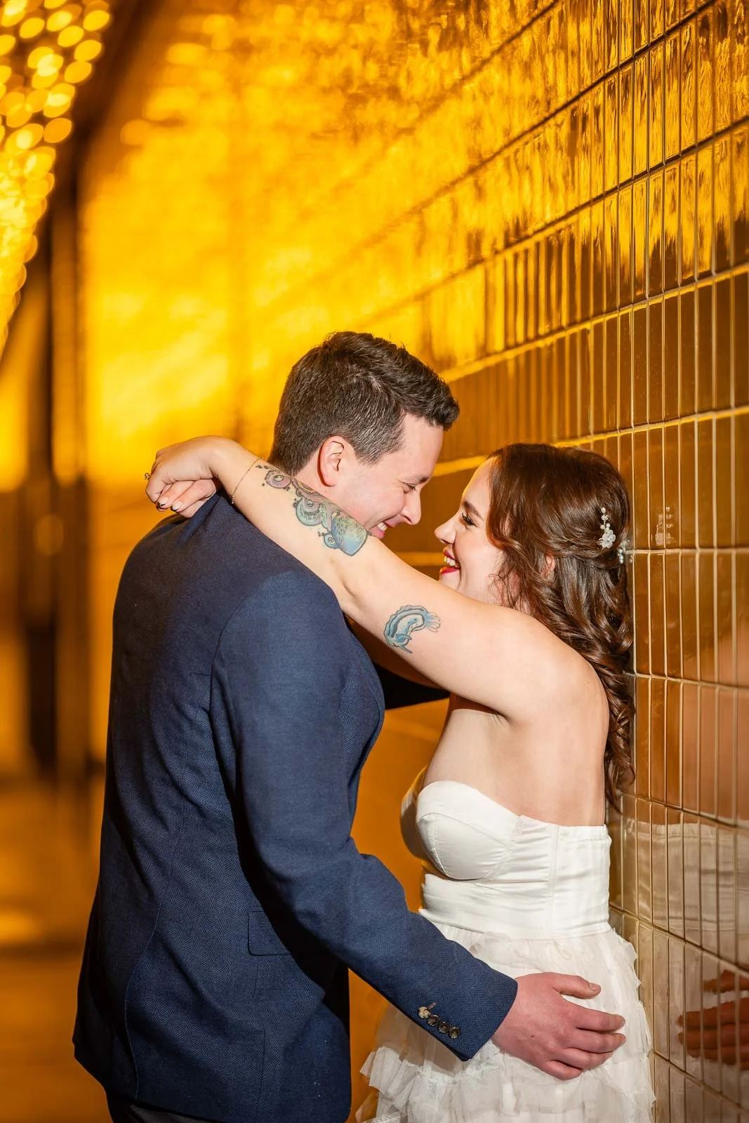 A couple dressed in wedding attire sharing an intimate moment against a yellow brick wall, smiling with their arms around each other.