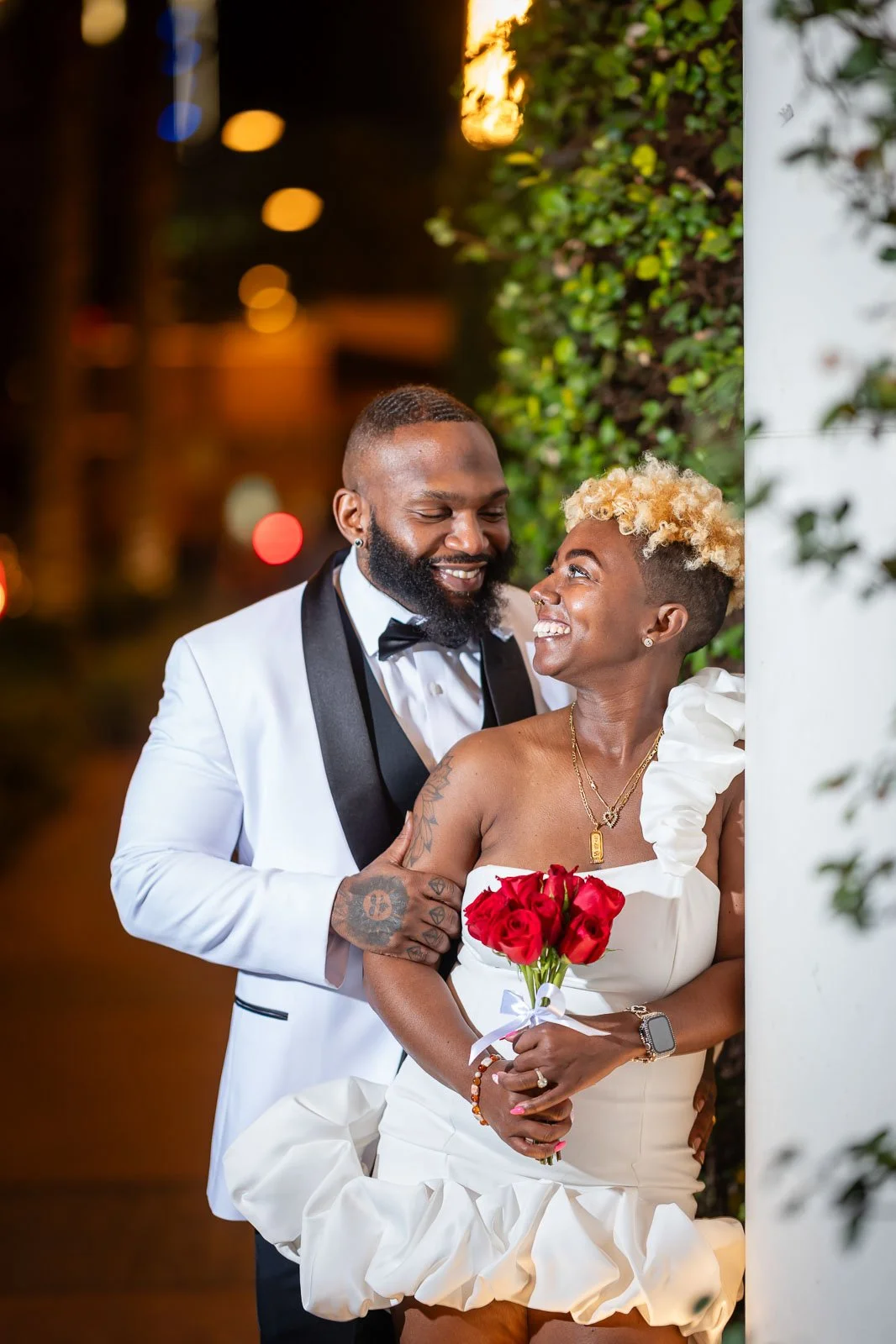 A happy couple dressed in wedding attire, smiling and looking at each other, with the woman holding a bouquet of red roses, standing outside on a city street at night.