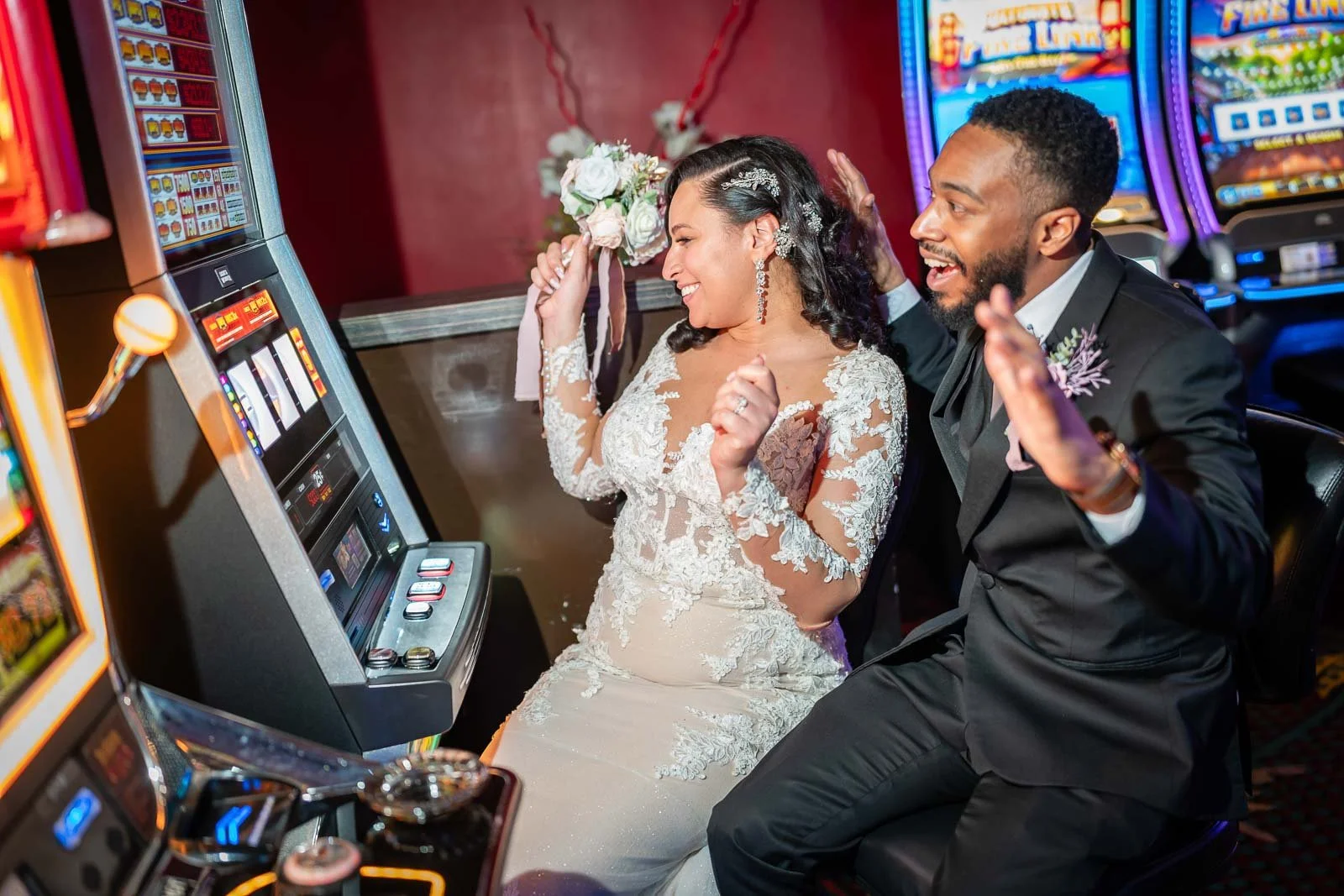 A bride and groom playing a slot machine in a casino. The bride is holding a bouquet and wearing a lace wedding dress, and the groom is in a black suit. They are laughing and enjoying their time together.