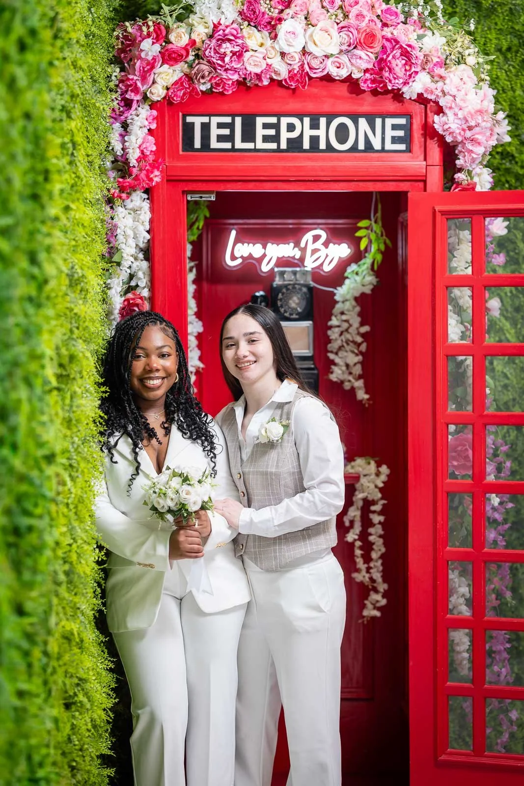Two women in white suits with boutonnieres and a bouquet of white flowers, smiling in front of a red phone booth decorated with pink and white flowers and a neon sign that says "Love you, Bye."