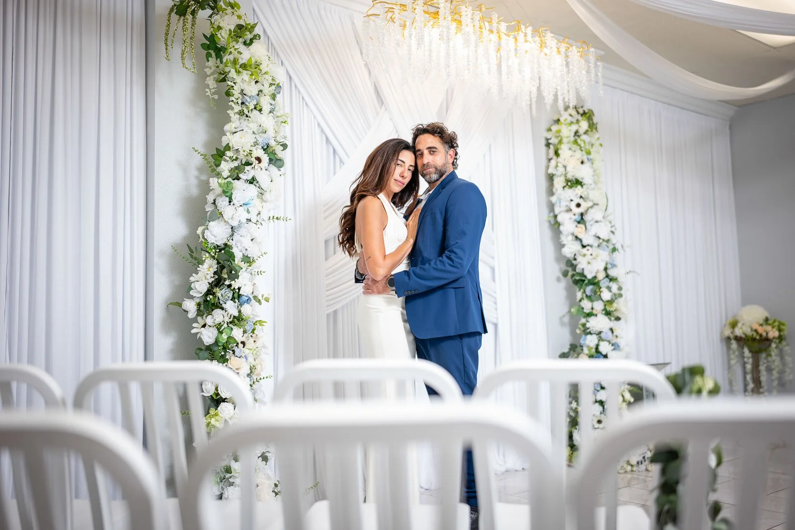 A couple dressed in wedding attire standing close together at a wedding ceremony, surrounded by white floral decorations and drapery.