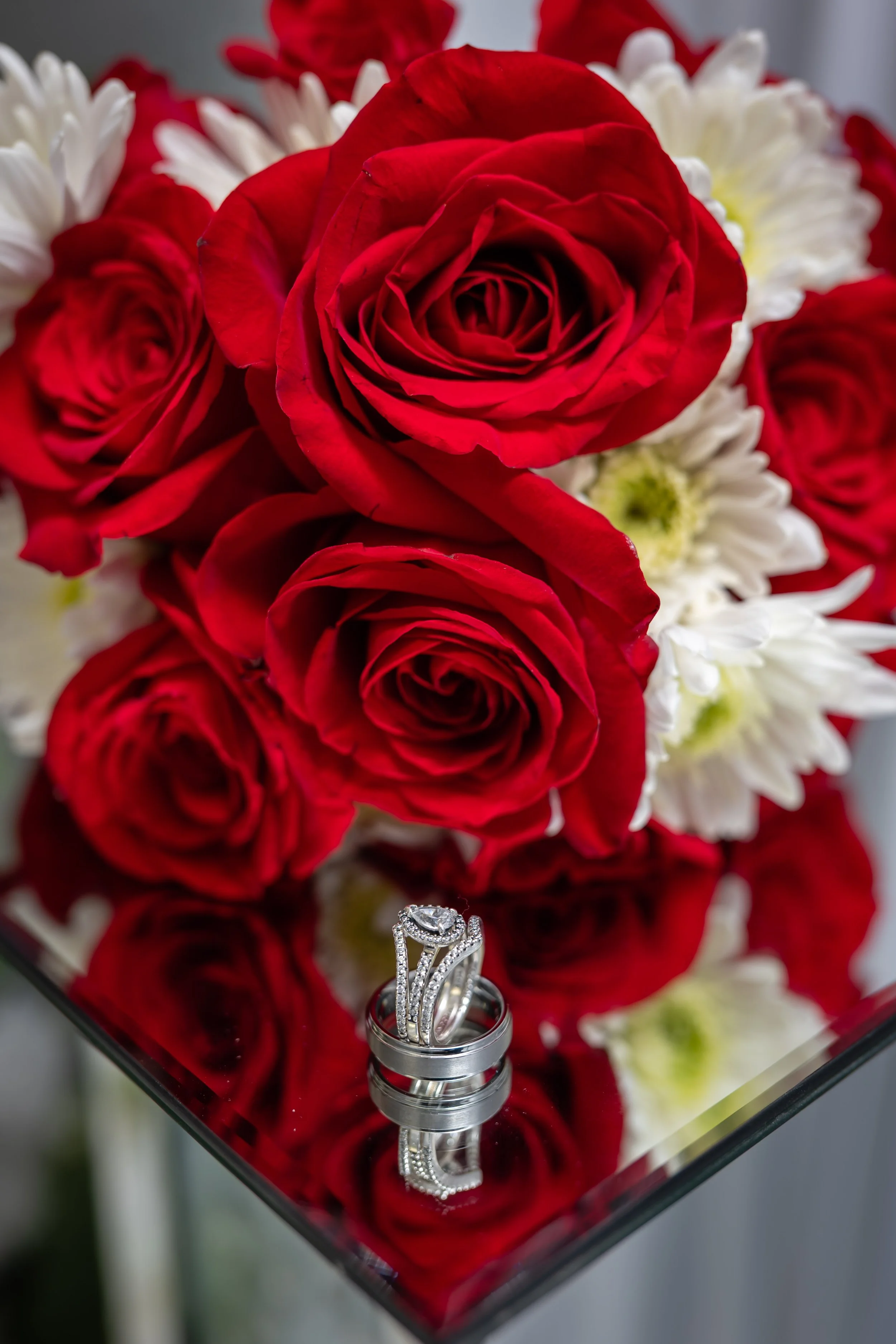 A bouquet of red roses and white daisies with a diamond engagement ring and wedding band placed on a reflective black surface.
