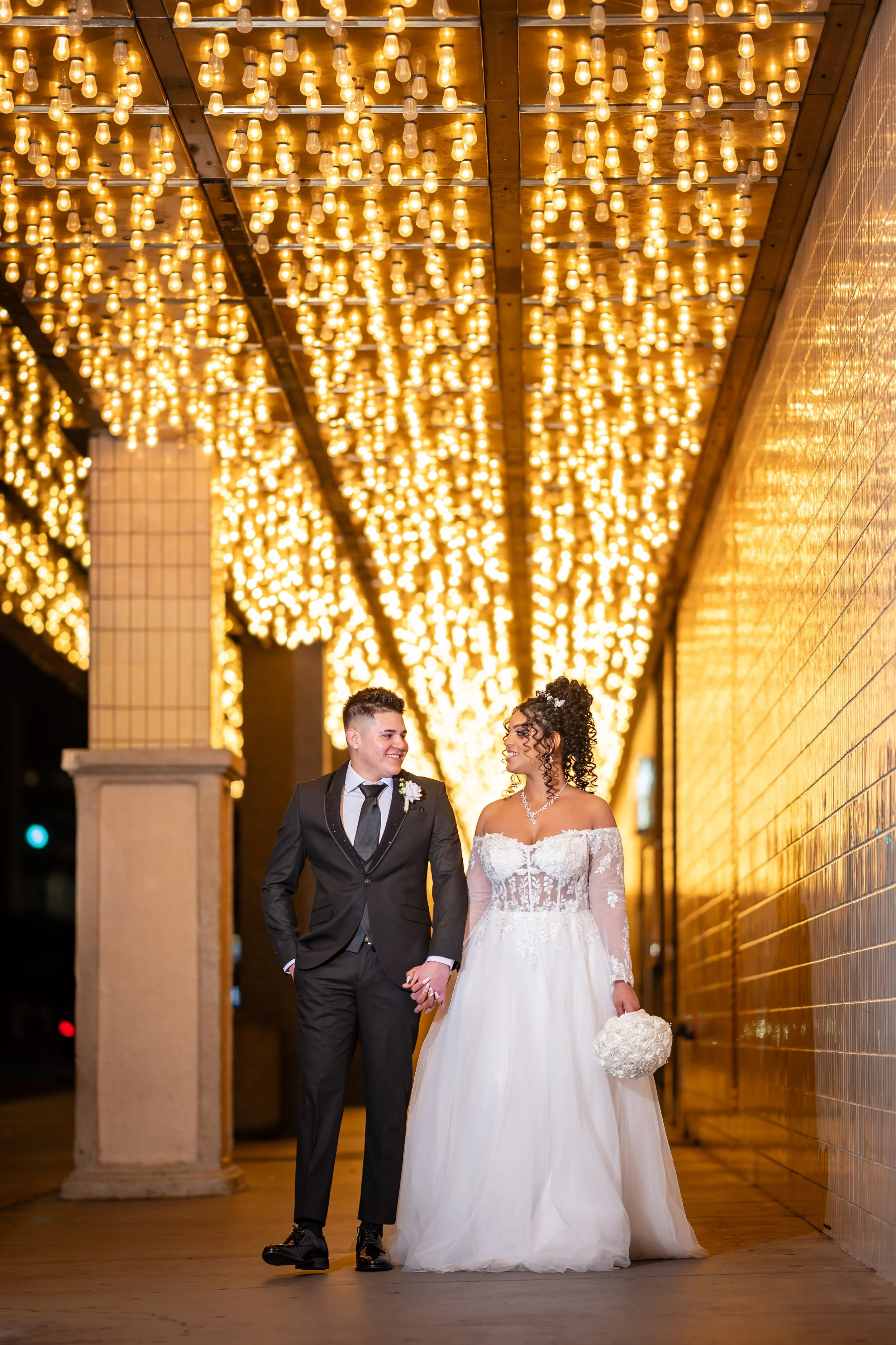 A newlywed couple walking under a canopy of warm string lights, with the bride holding a bouquet and wearing a lace wedding dress, and the groom in a black suit and tie.