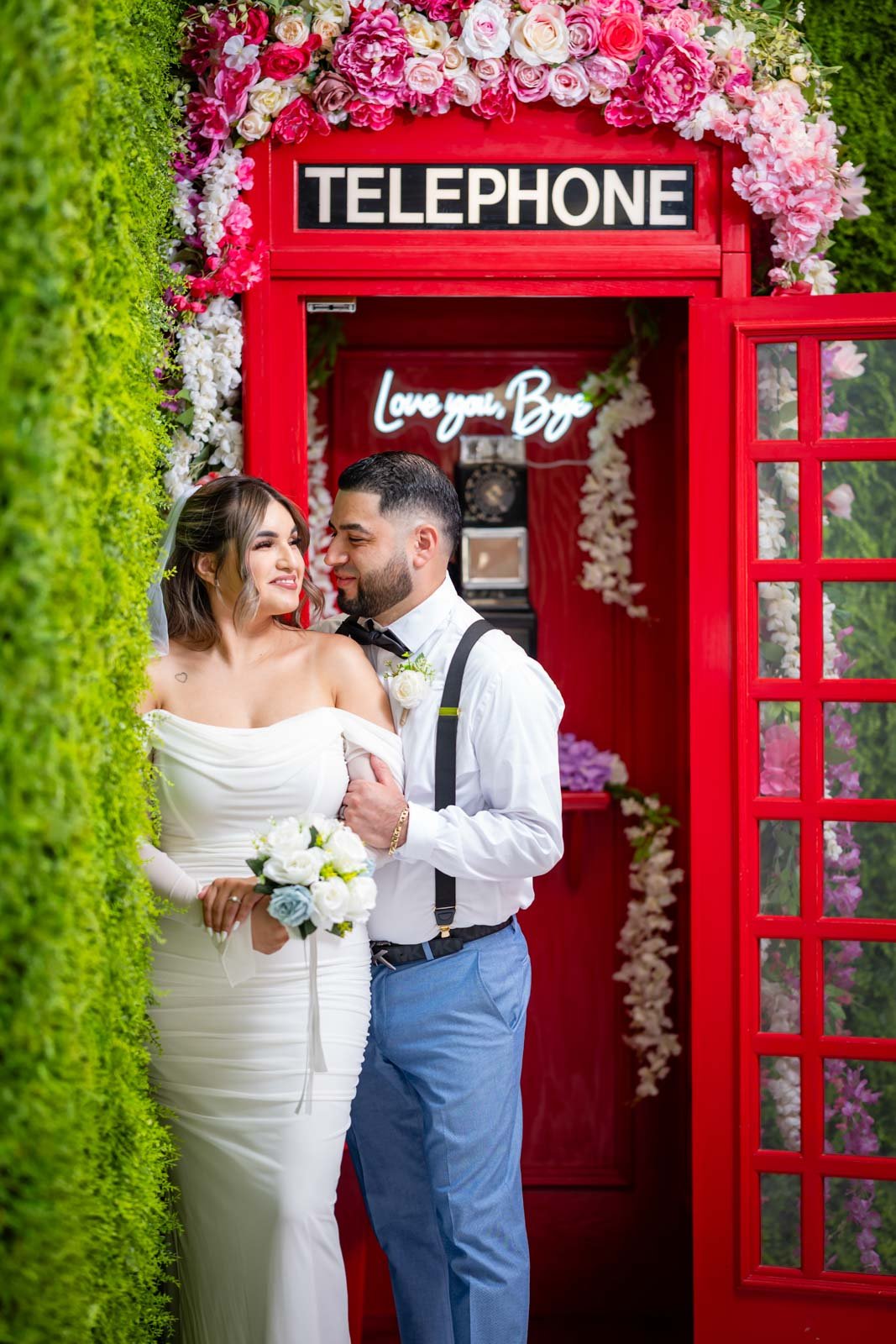A bride and groom standing in front of a vintage red telephone booth decorated with pink and white flowers, with a neon sign inside reading 'Love you, Bro'.