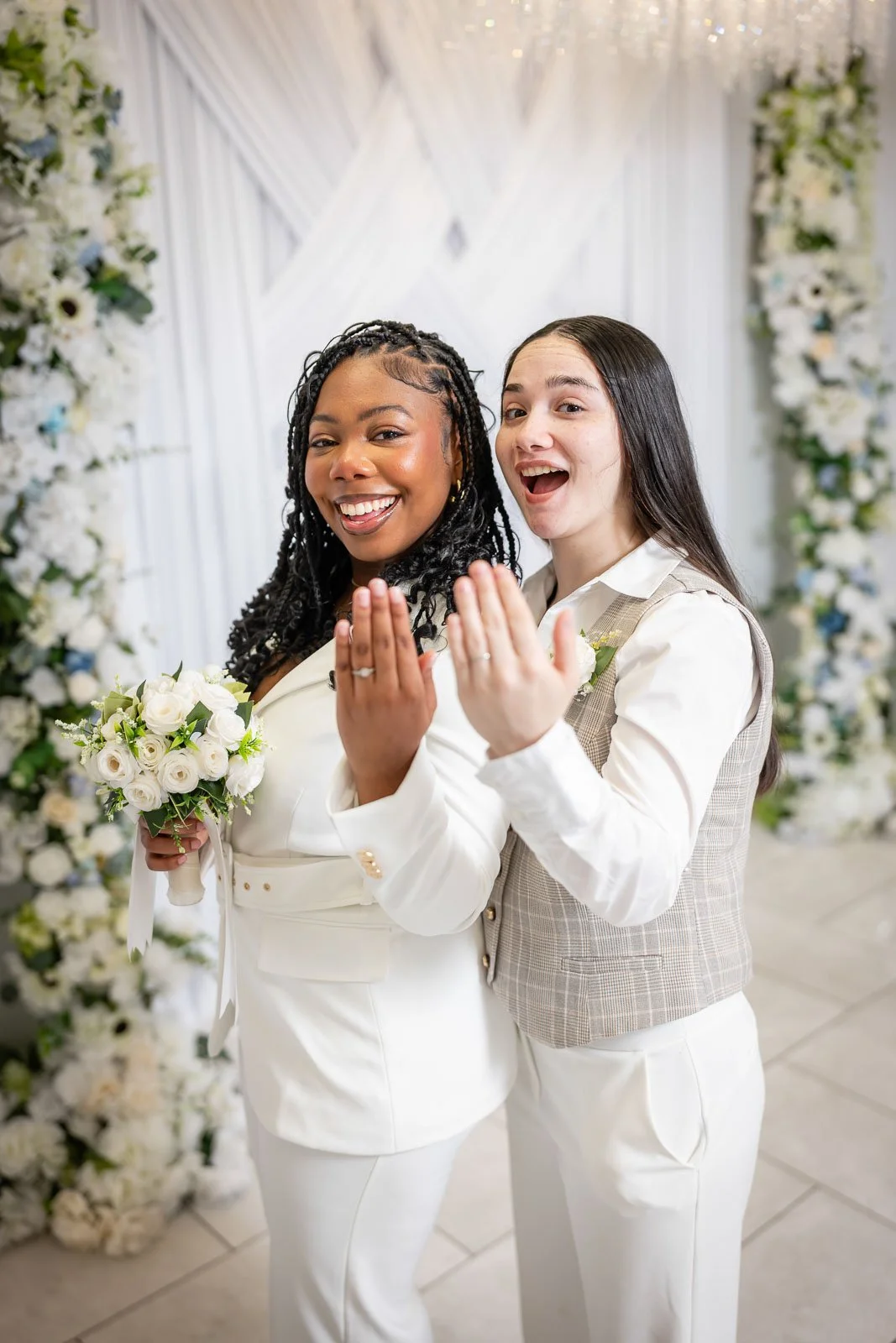 Two women at the Say I Do Wedding Chapel, showing off matching wedding rings and smiling, standing in front of a floral backdrop.