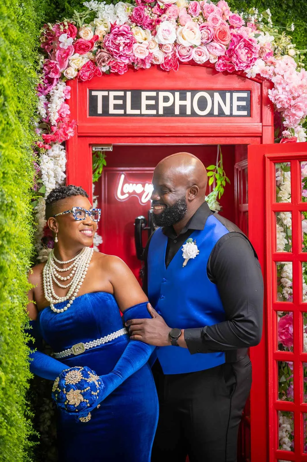 A smiling woman in a royal blue dress and pearl jewelry posing with a smiling man in a blue vest and black shirt inside a red British-style phone booth decorated with pink and white flowers.