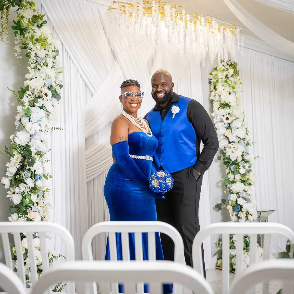 A couple dressed in matching blue attire stands together at a wedding venue with white floral decorations and draped fabric backdrop.